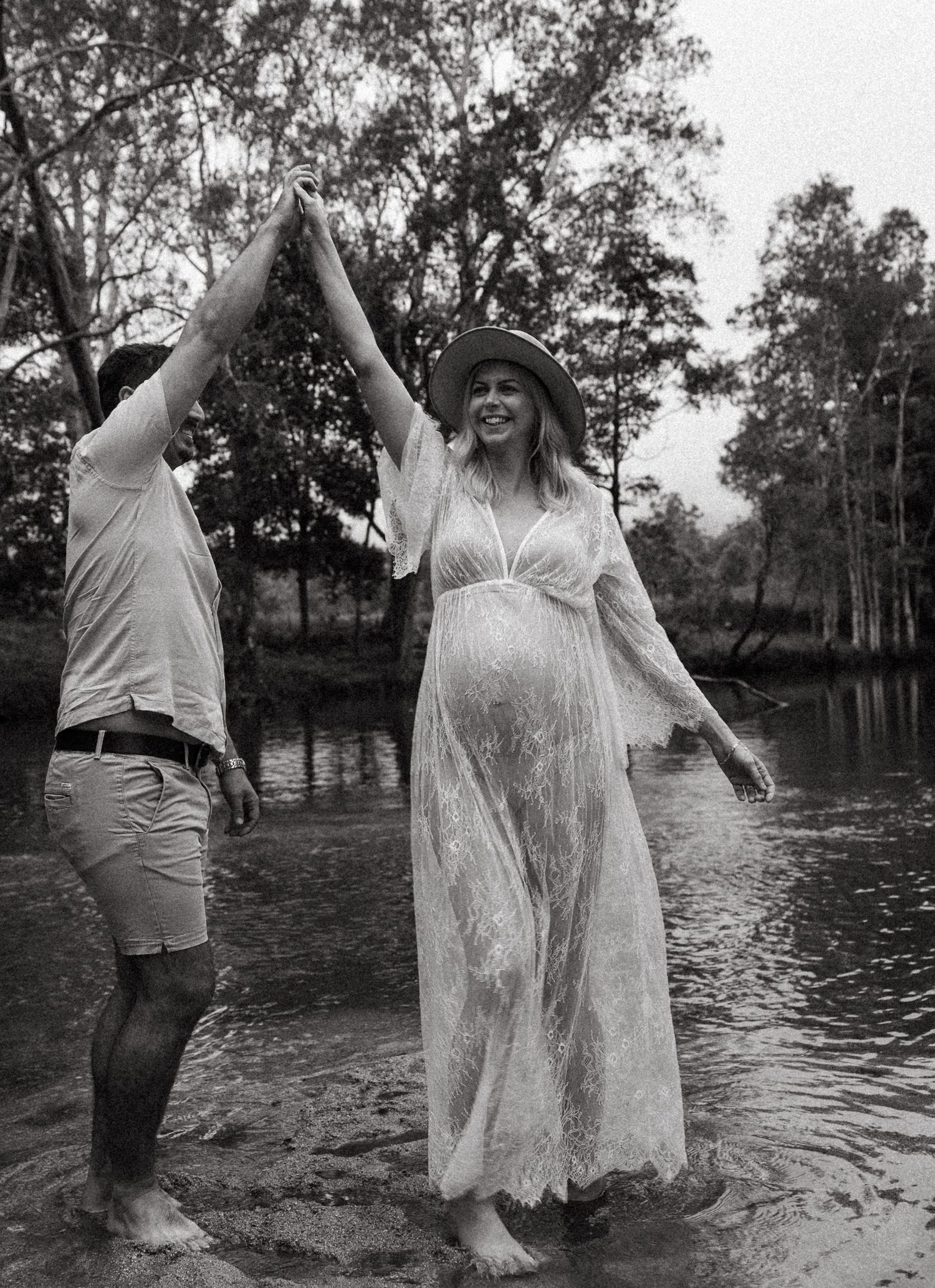 A couple stands in shallow water, with the pregnant person wearing a flowing white lace dress amidst a scenic lake backdrop.