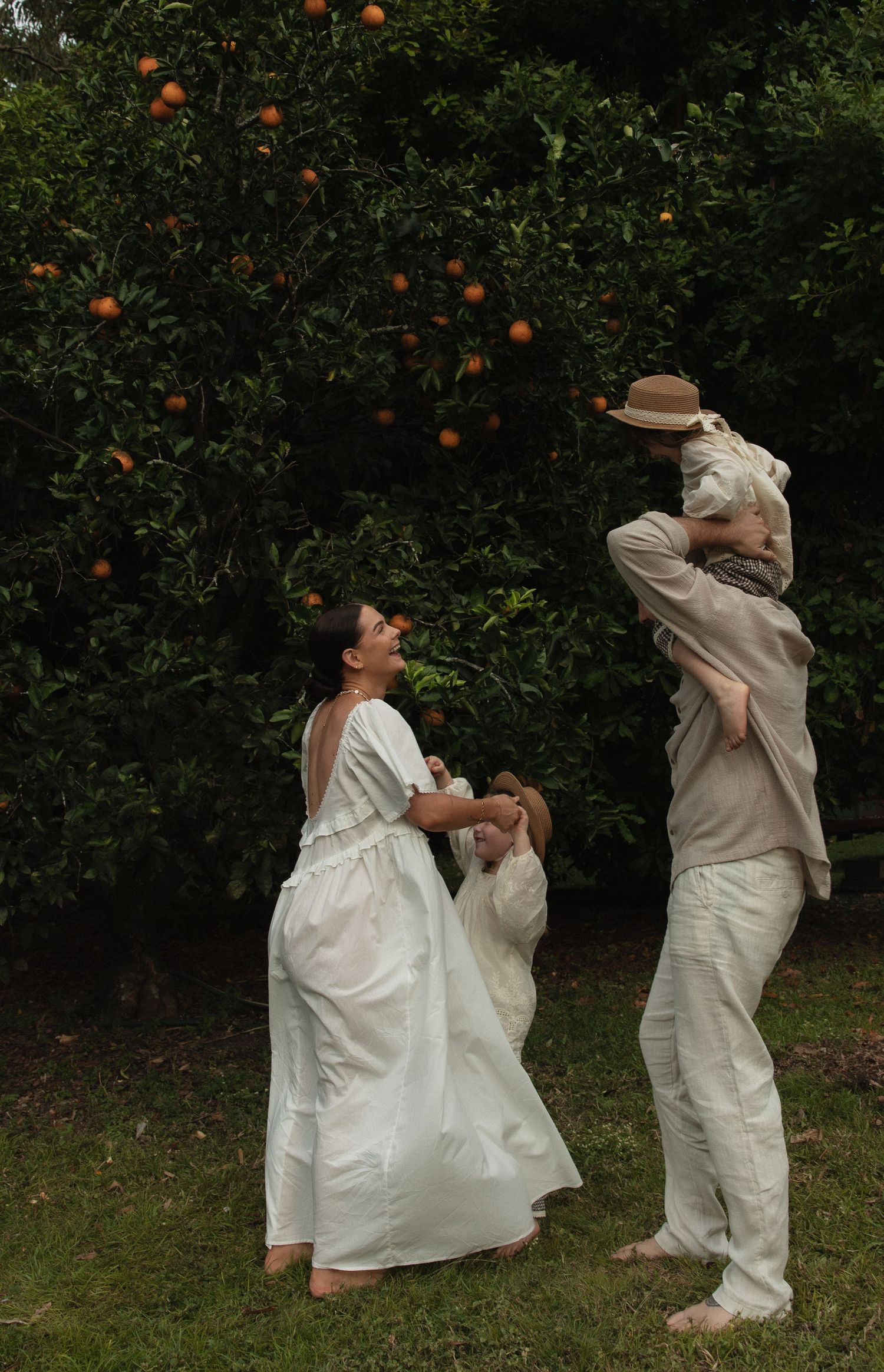 People in flowing white outfits playfully reach for oranges in a verdant citrus grove on a sunny day.