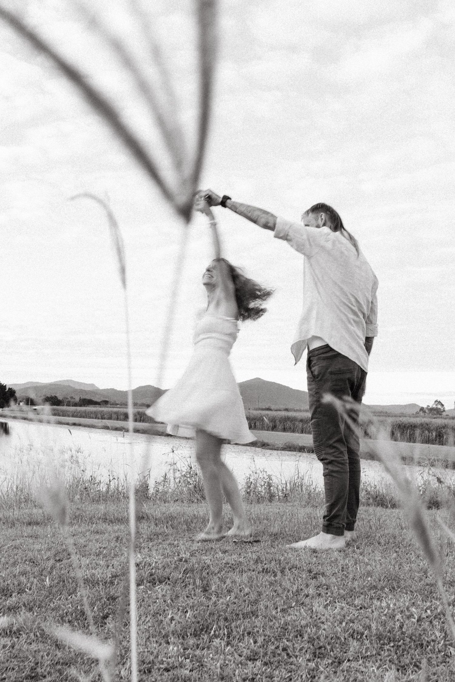 A couple dances joyfully in a field with mountains in the background, captured in black and white photography.