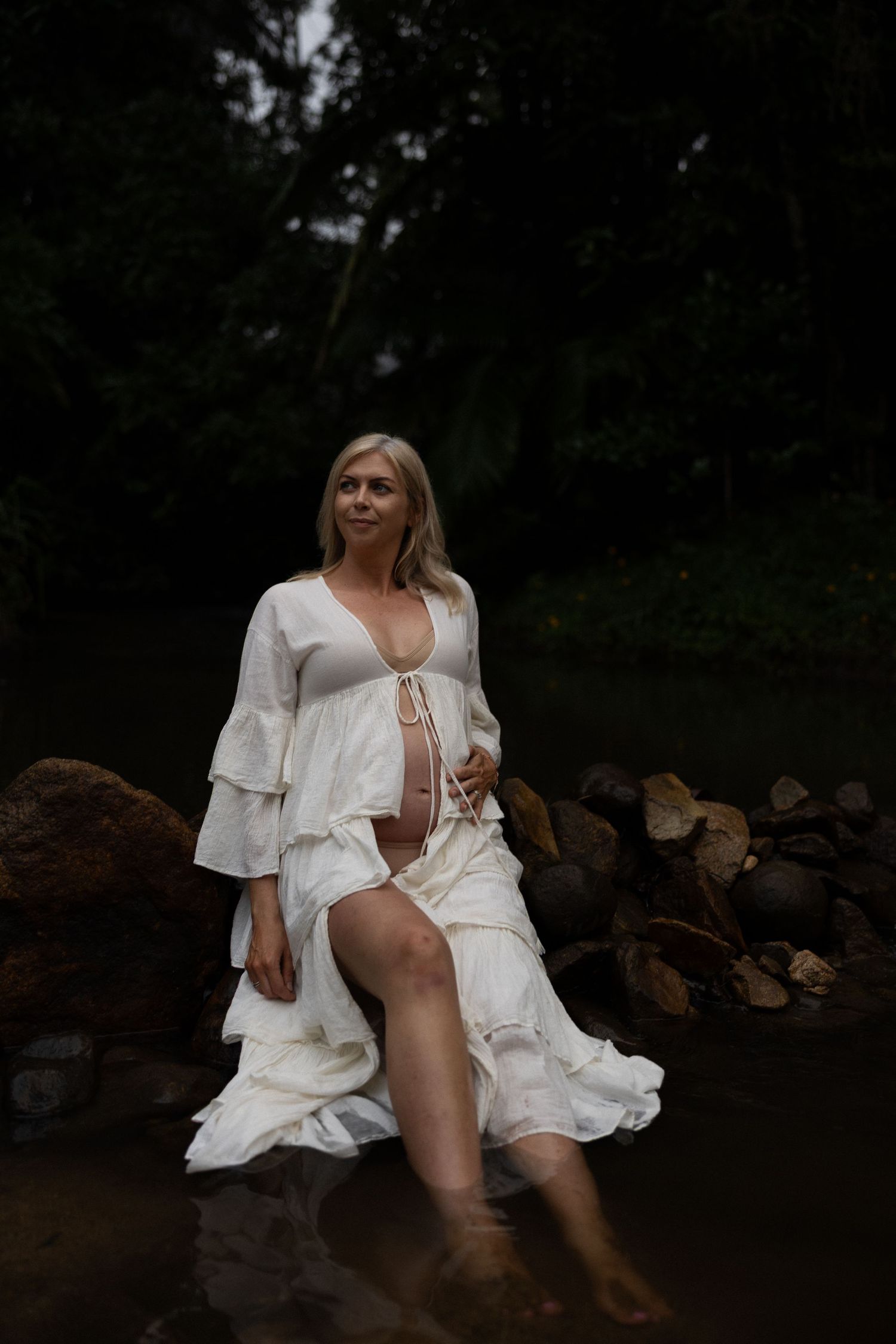 A woman in a white flowing dress sits at the edge of a pond with stacked logs nearby in a moody forest setting.