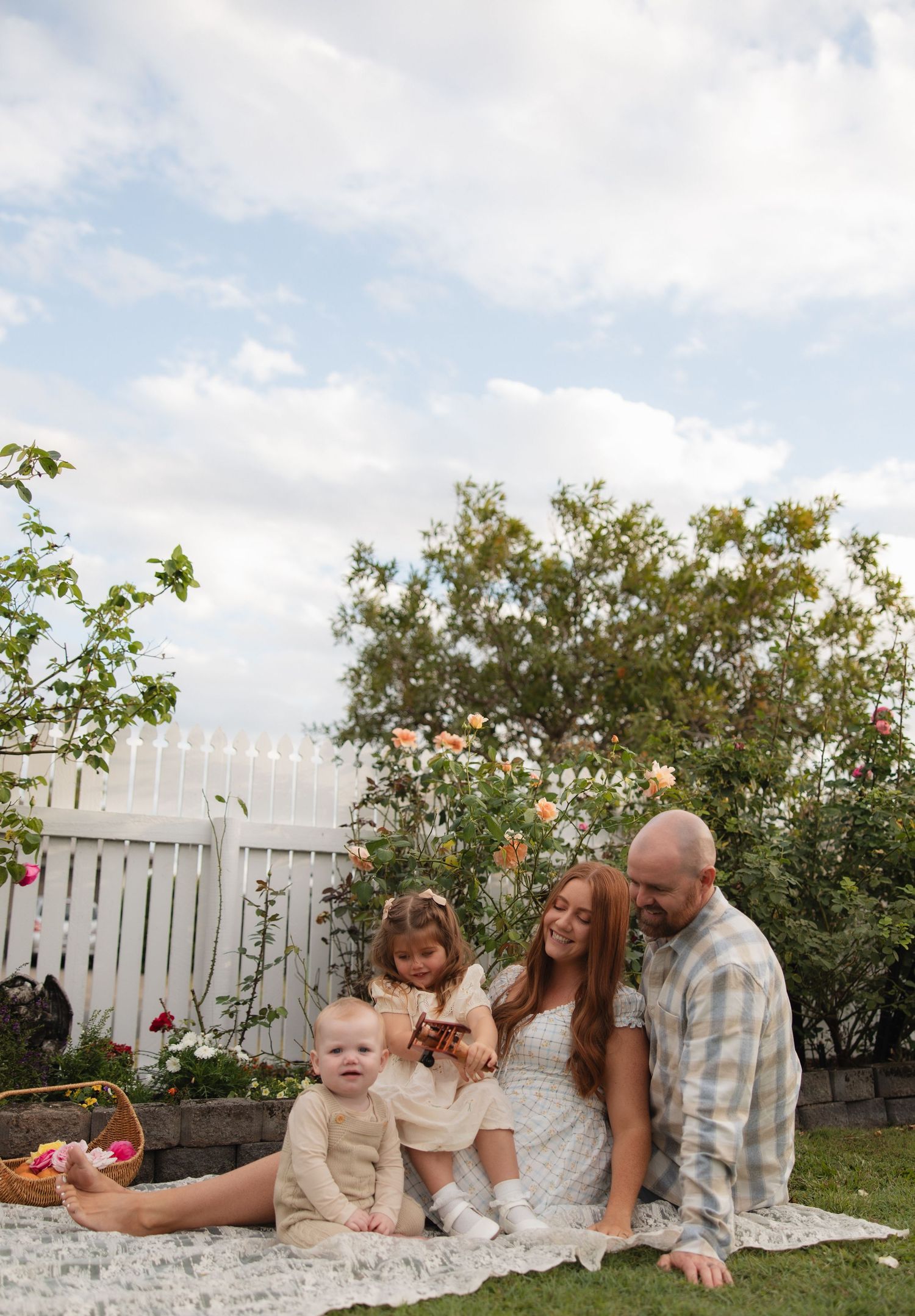 Family enjoying a picnic on a white blanket in their backyard garden with roses and a white fence on a sunny day.