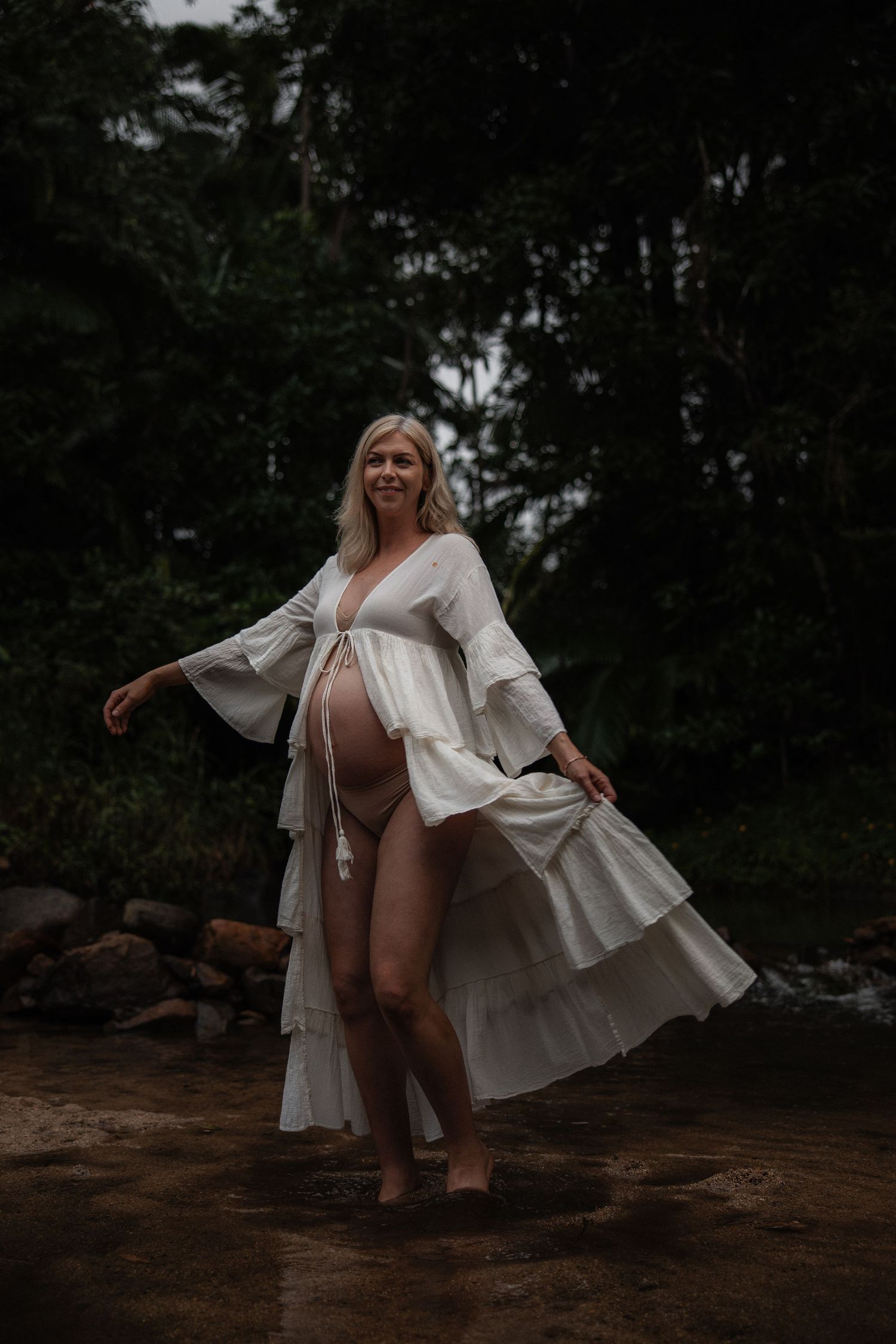 A figure in a flowing white tiered dress poses gracefully by a stream in a moody forest setting.