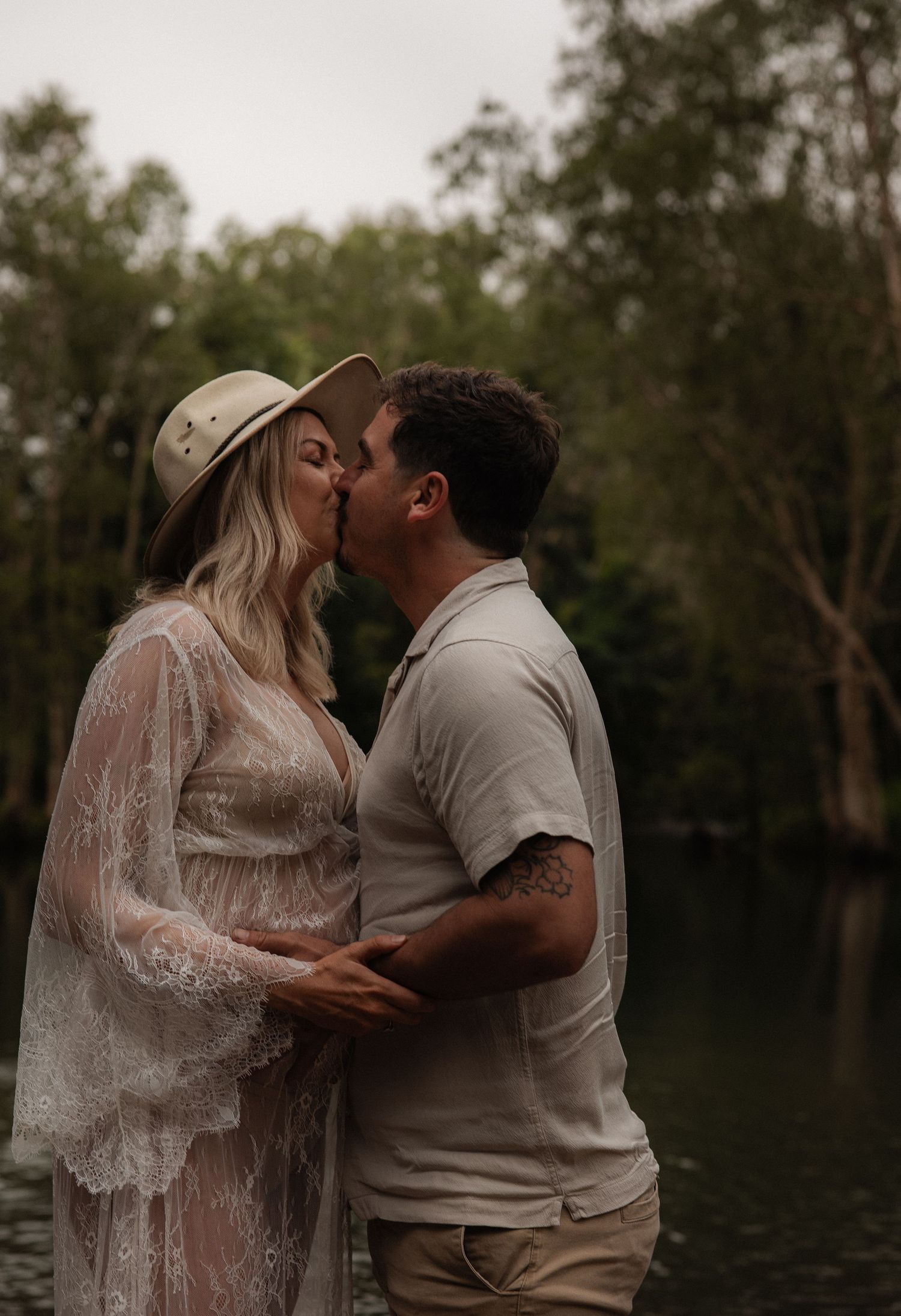 A romantic couple shares a tender kiss by a serene lake, with the bride wearing a flowing lace dress against a wooded backdrop.