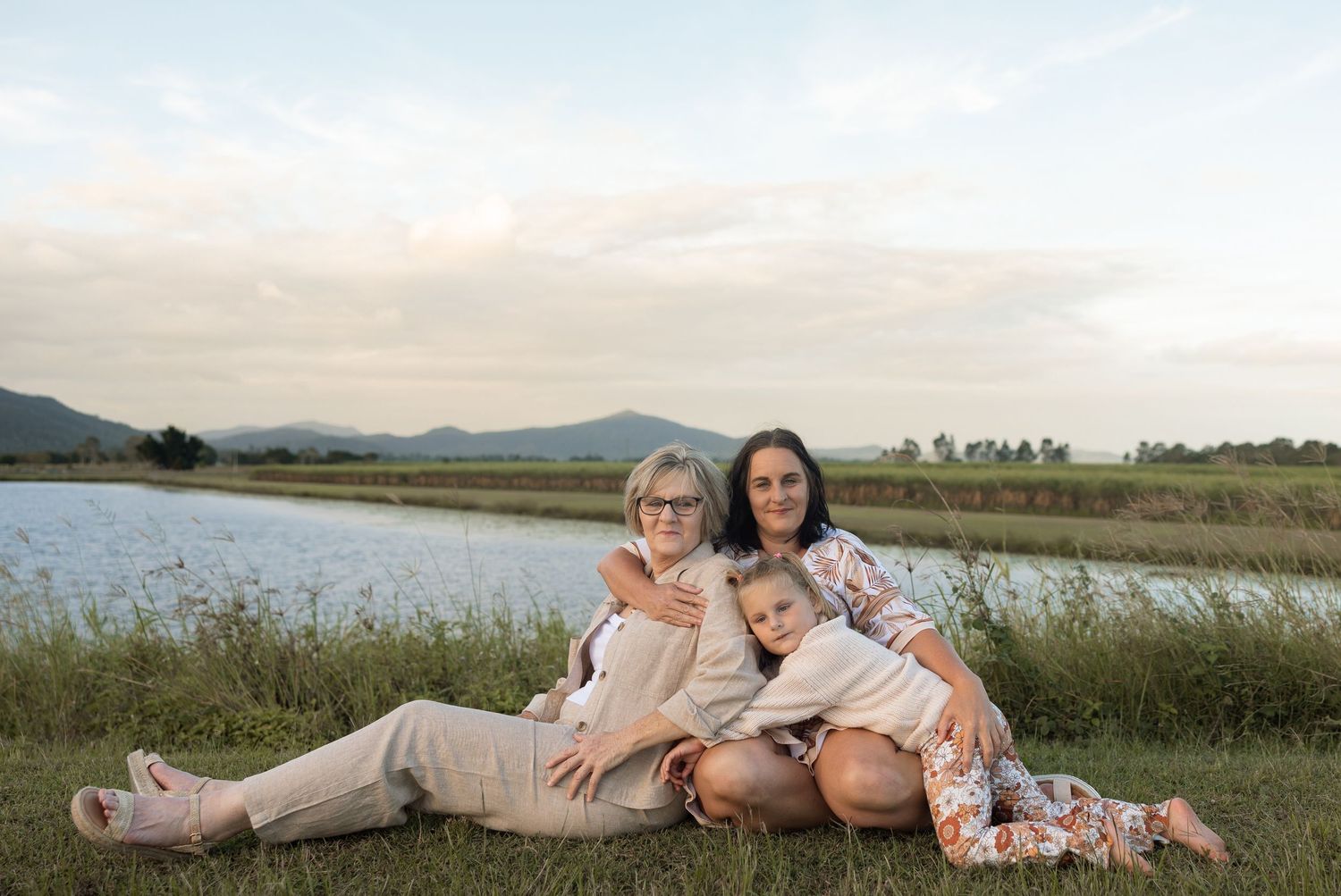 Family relaxing together on grassy lakeside in coordinated neutral and floral outfits.
