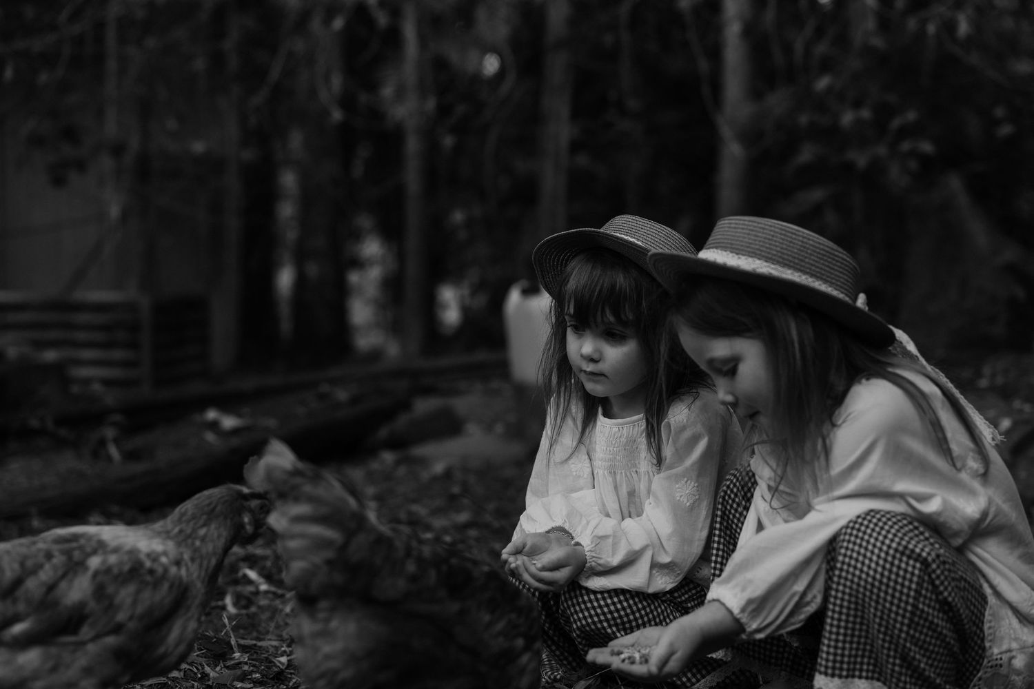 Two children in summer dresses and hats sit together outdoors in a moody black and white photo.
