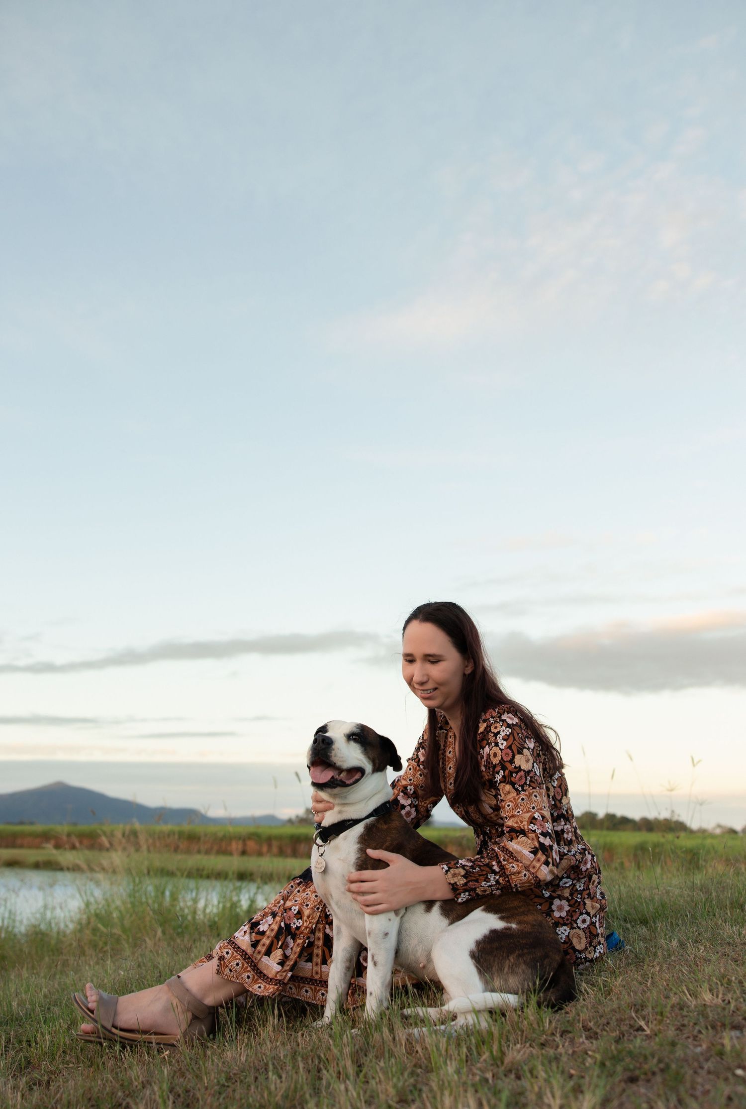 A person in a floral dress sits on grass at sunset with their black and white Border Collie dog near a lake.