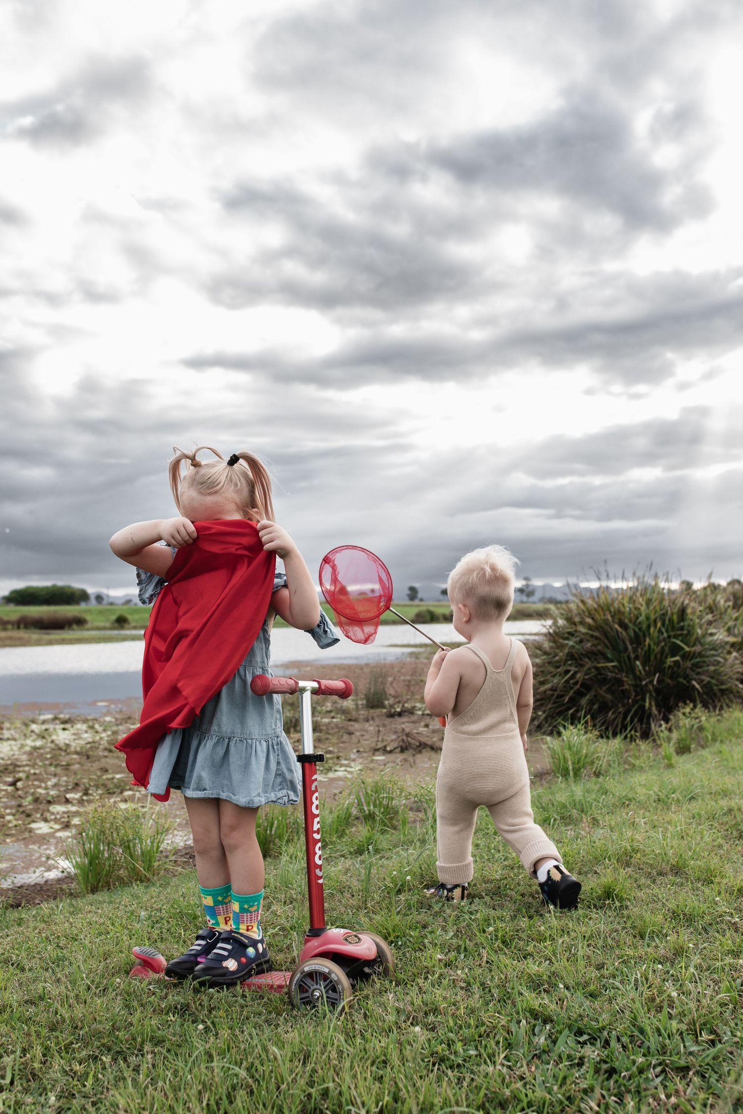 Children playing outdoors with a red scooter and butterfly net against a dramatic cloudy sky.