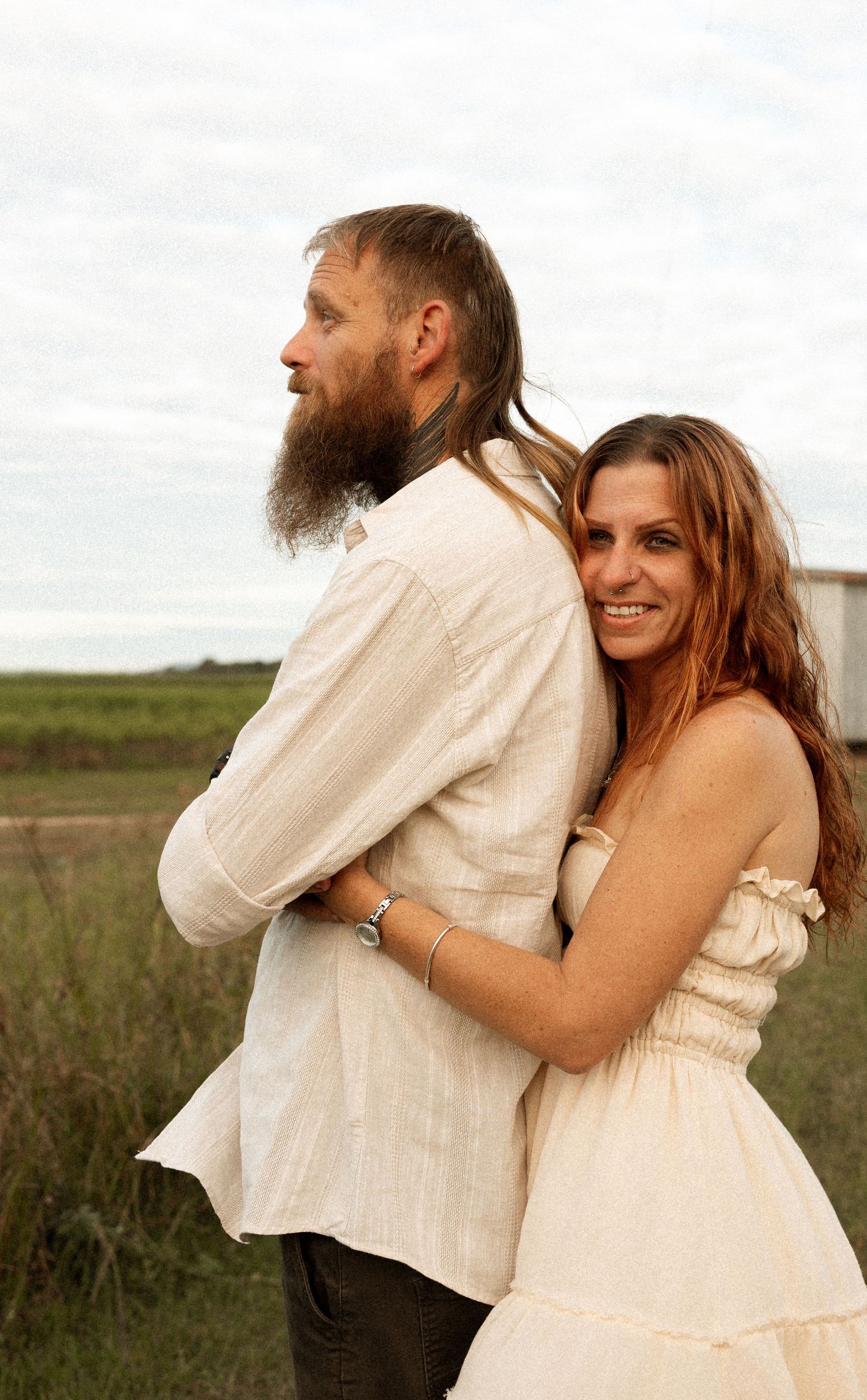 A couple embraces in a grassy field at sunset, with her in a white dress hugging him from behind as he wears a light shirt.