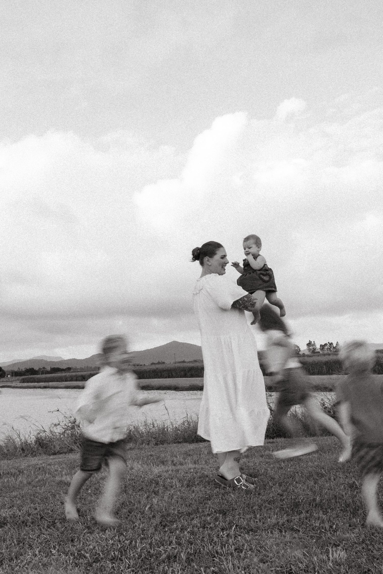 Black and white scene of children playing around figure in white dress by the water.