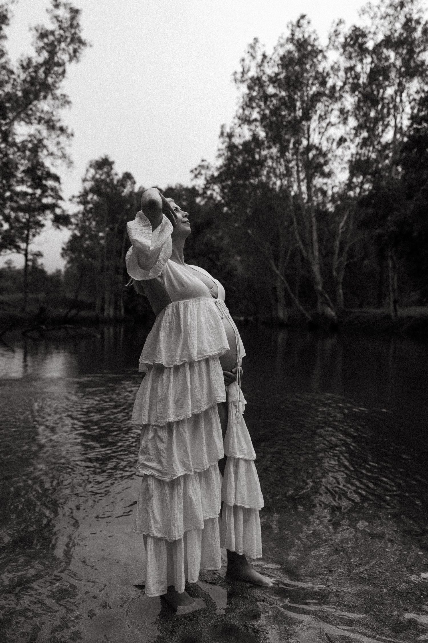 Figure in flowing white tiered dress standing in dark water surrounded by trees in moody black and white photography.