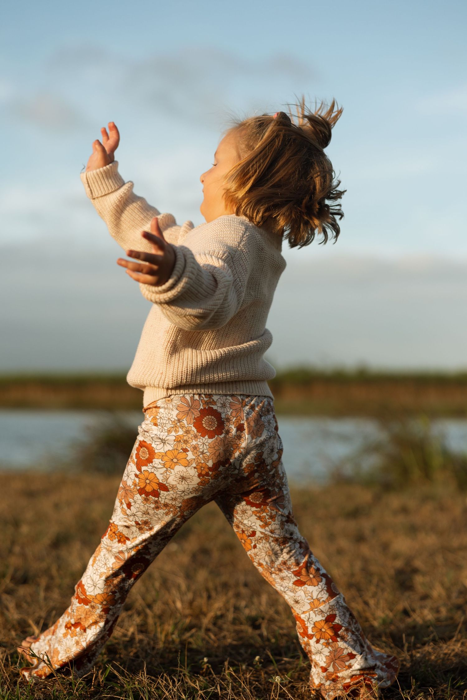 Child in beige sweater and floral pants dances joyfully against a waterfront landscape at sunset.