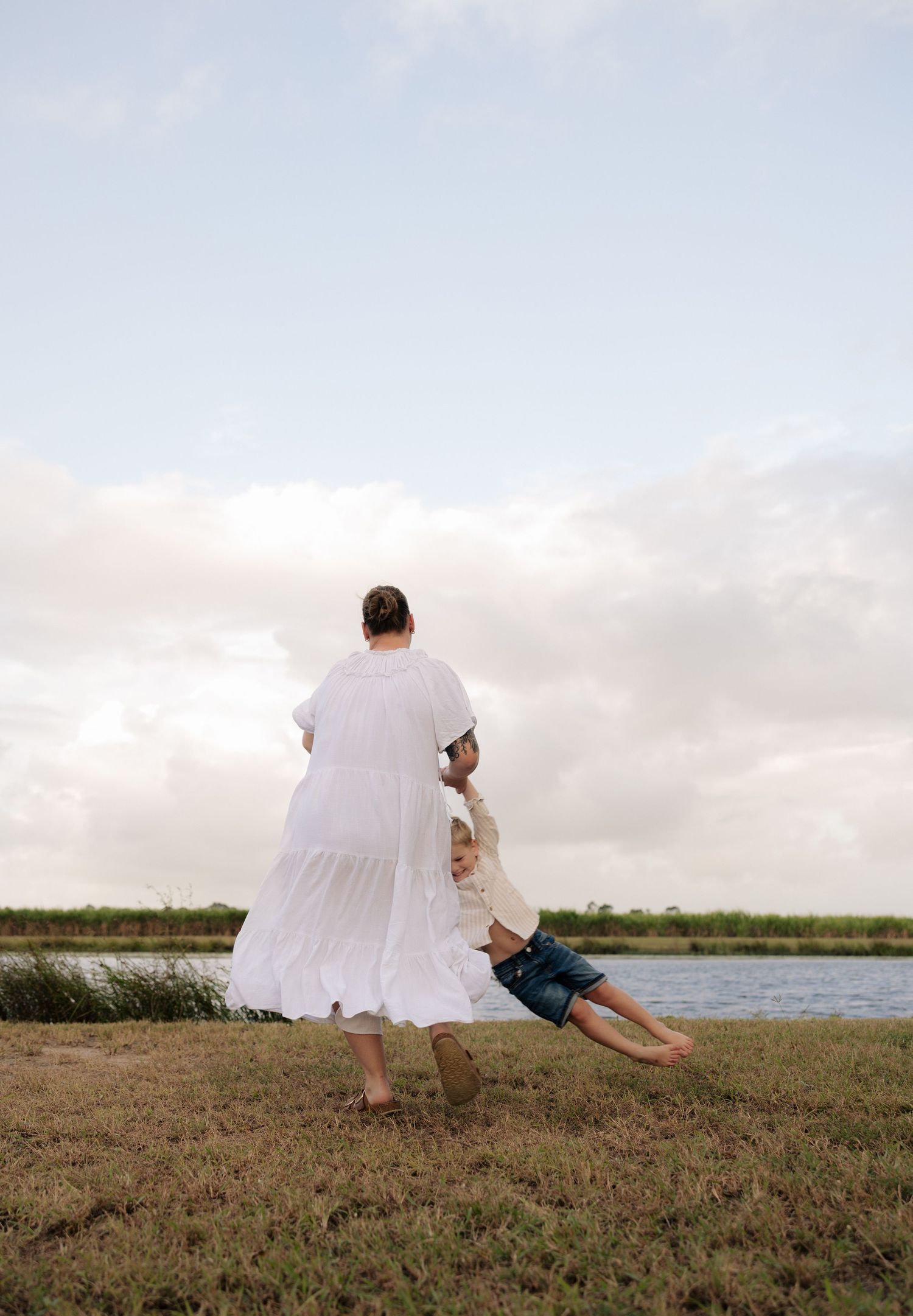 Someone in a white dress swings a child by the waterside at sunset.