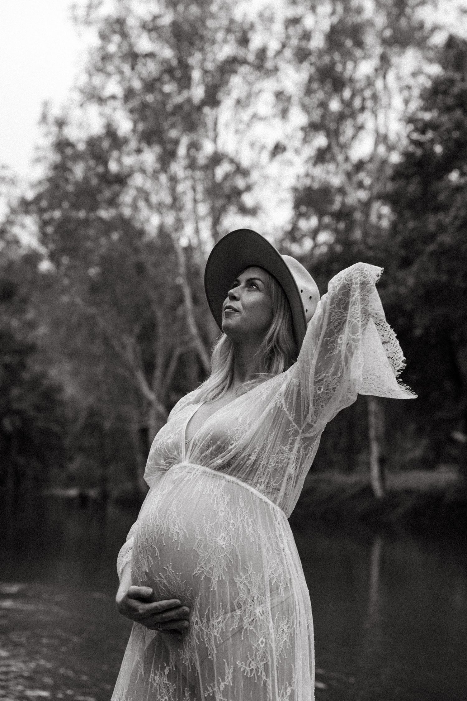 A pregnant woman in a sheer white lace dress and hat poses by a lake in a dreamy black and white photograph.