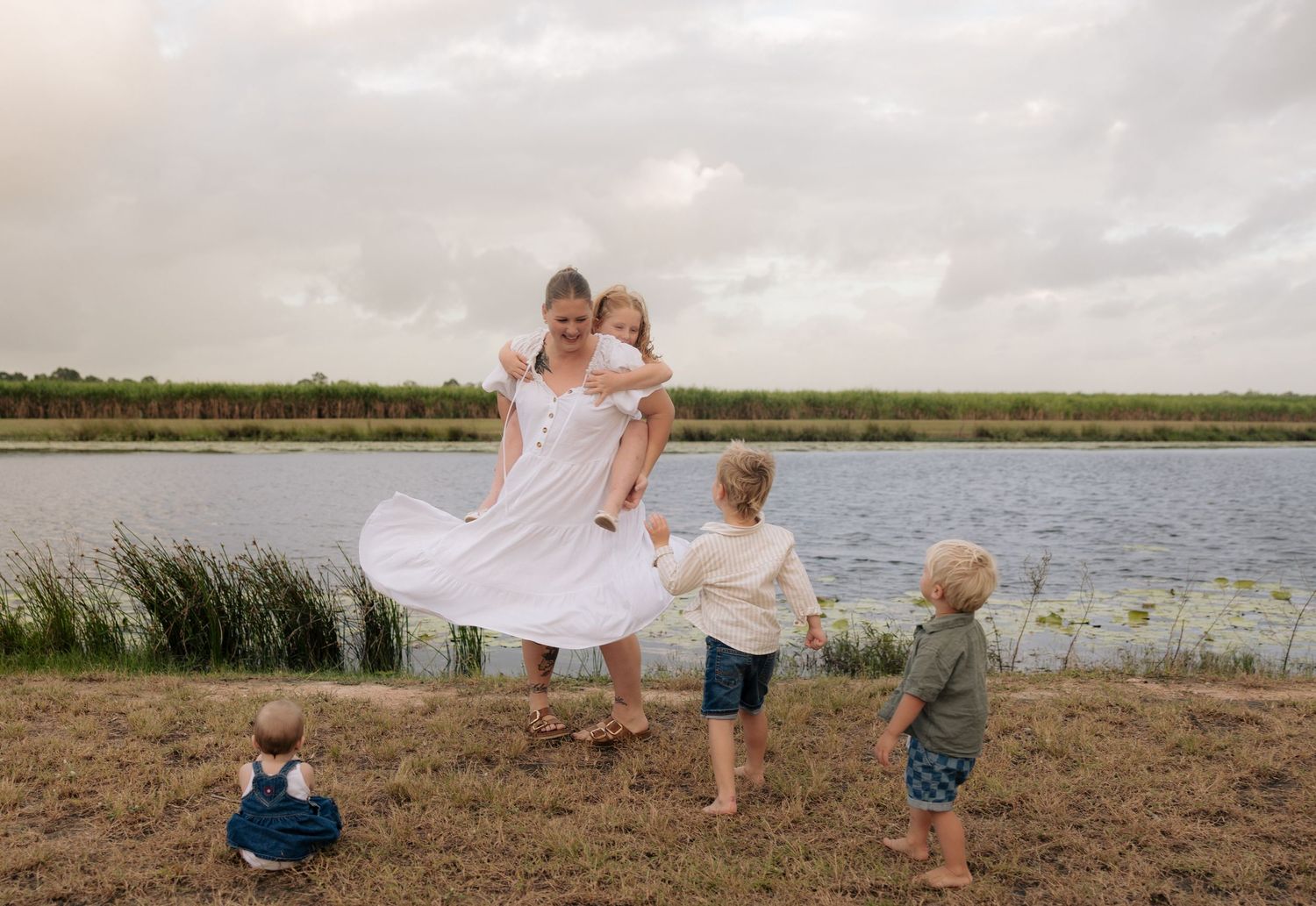 A family in white outfits plays together by a serene lake on a cloudy day.