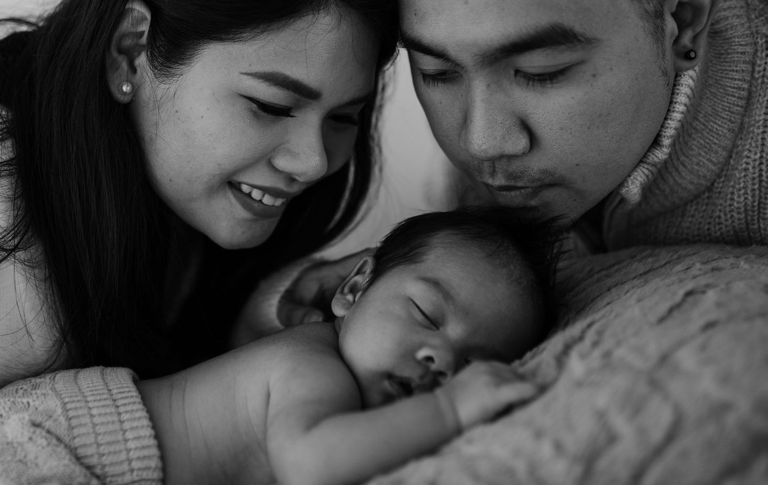 A black and white intimate portrait of a family gazing at their sleeping newborn.