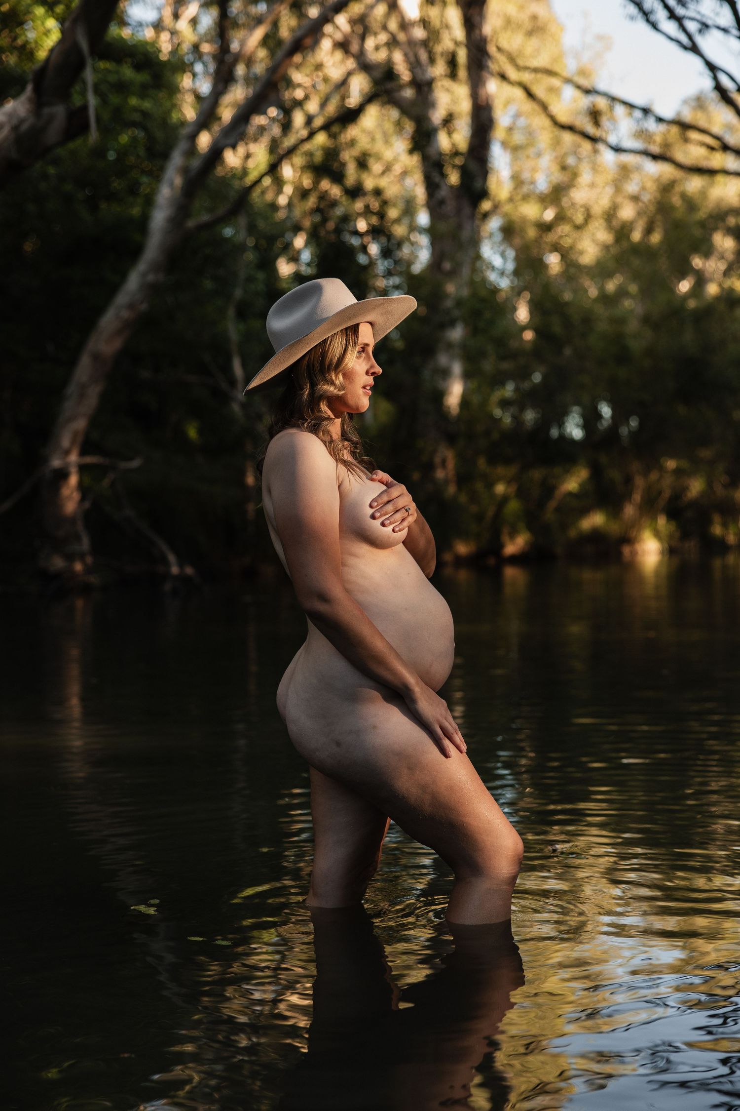 Maternity art photo of expecting mother standing in sunlit water wearing a wide-brimmed hat at sunset.
