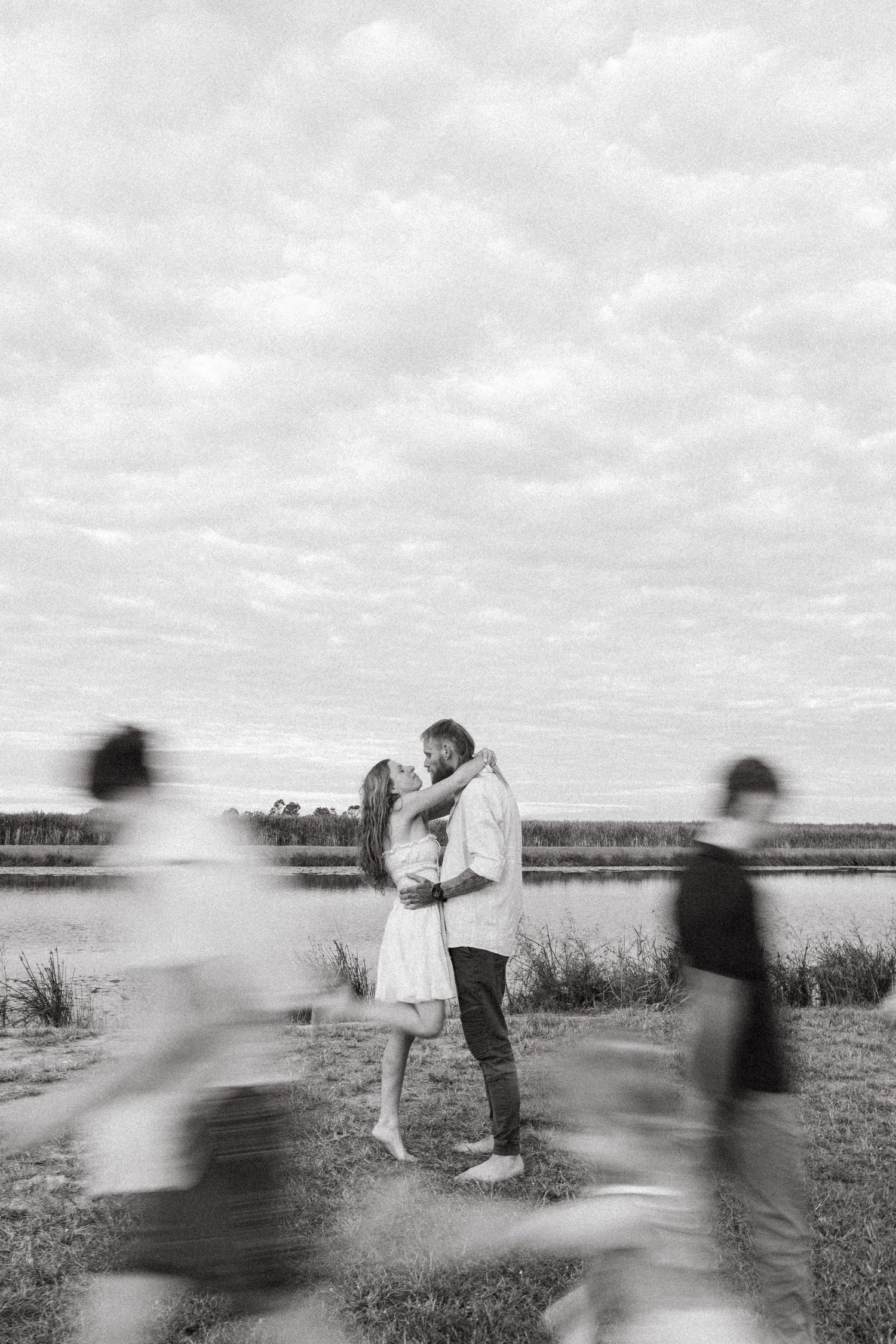 A romantic couple embraces by a waterfront while blurred figures pass by in black and white photography.