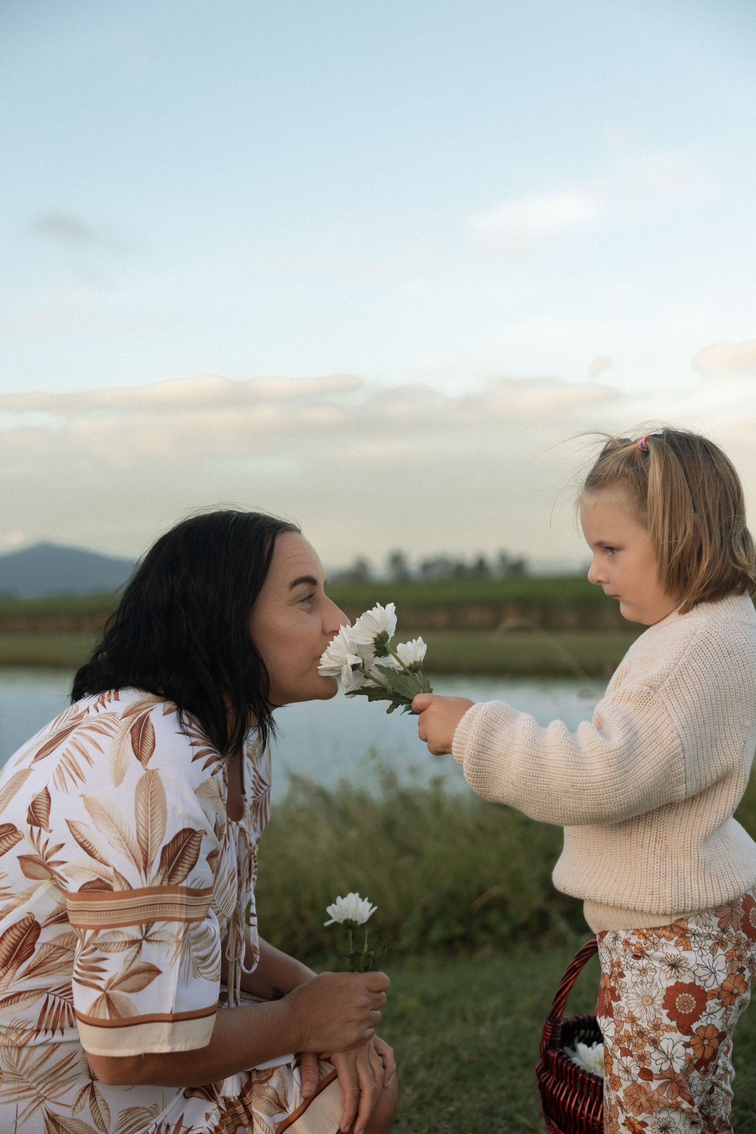 Sweet moment sharing wildflowers by the lake in coordinating cream and floral outfits.