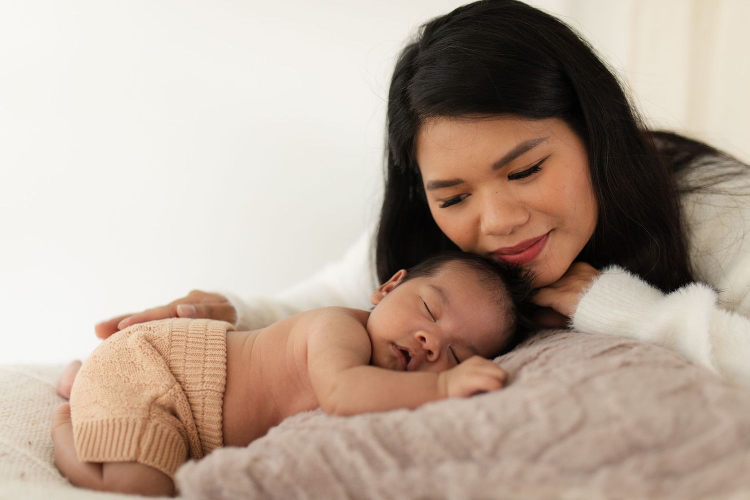 Sweet bonding moment with sleeping newborn in beige knit clothing on soft bedding.