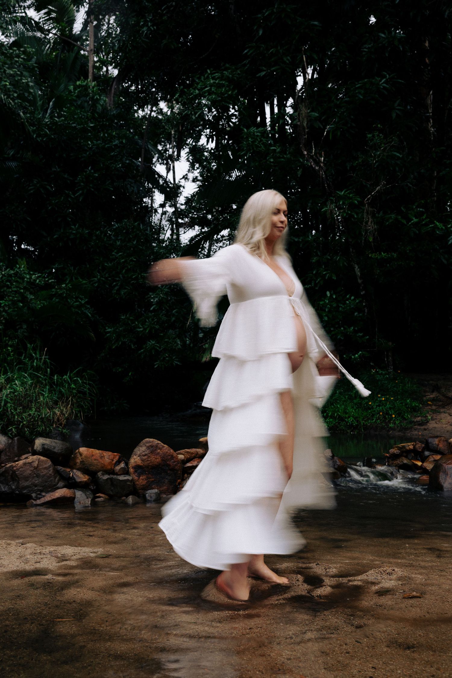 A figure in a flowing white tiered dress twirls gracefully by a rocky stream in a dark forest setting.