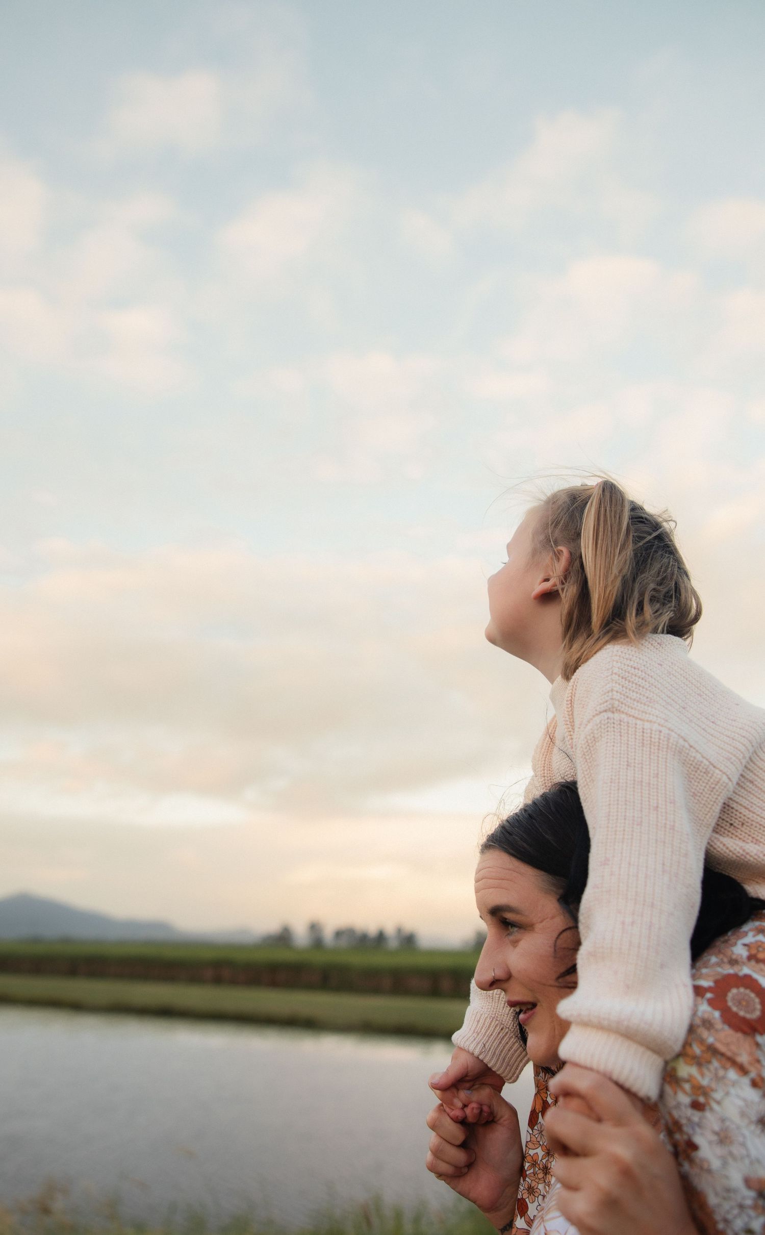 Parent carrying child on shoulders against sunset sky and lake view.