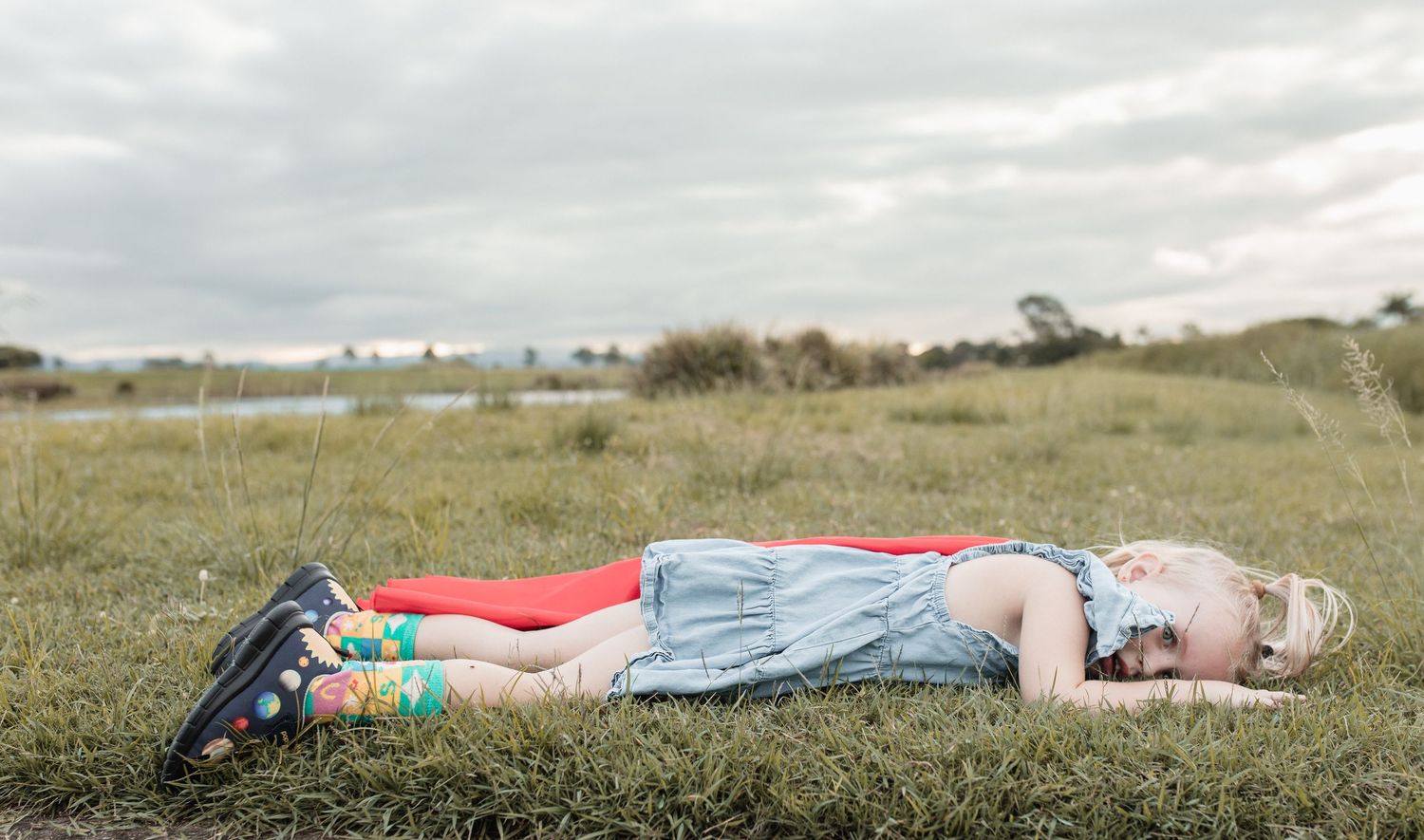 Child lying down in grass wearing red and denim clothing against cloudy sky.