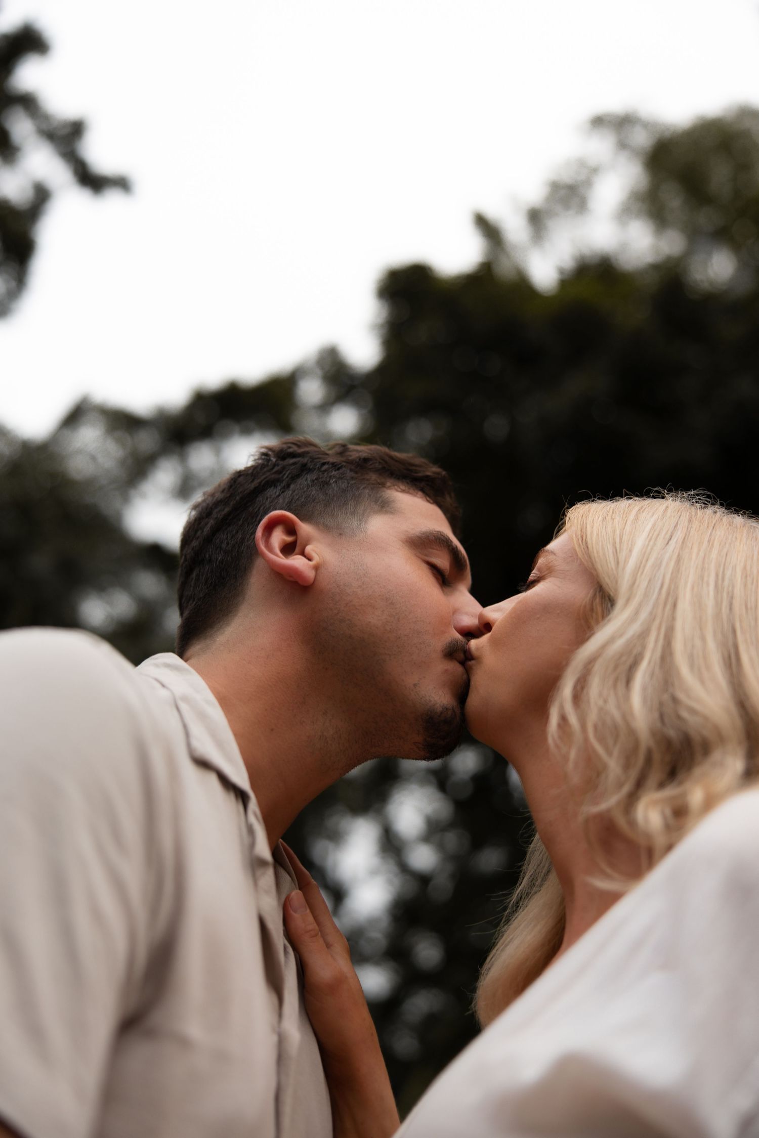 A romantic couple shares a tender embrace while kissing outdoors against a dark natural backdrop.