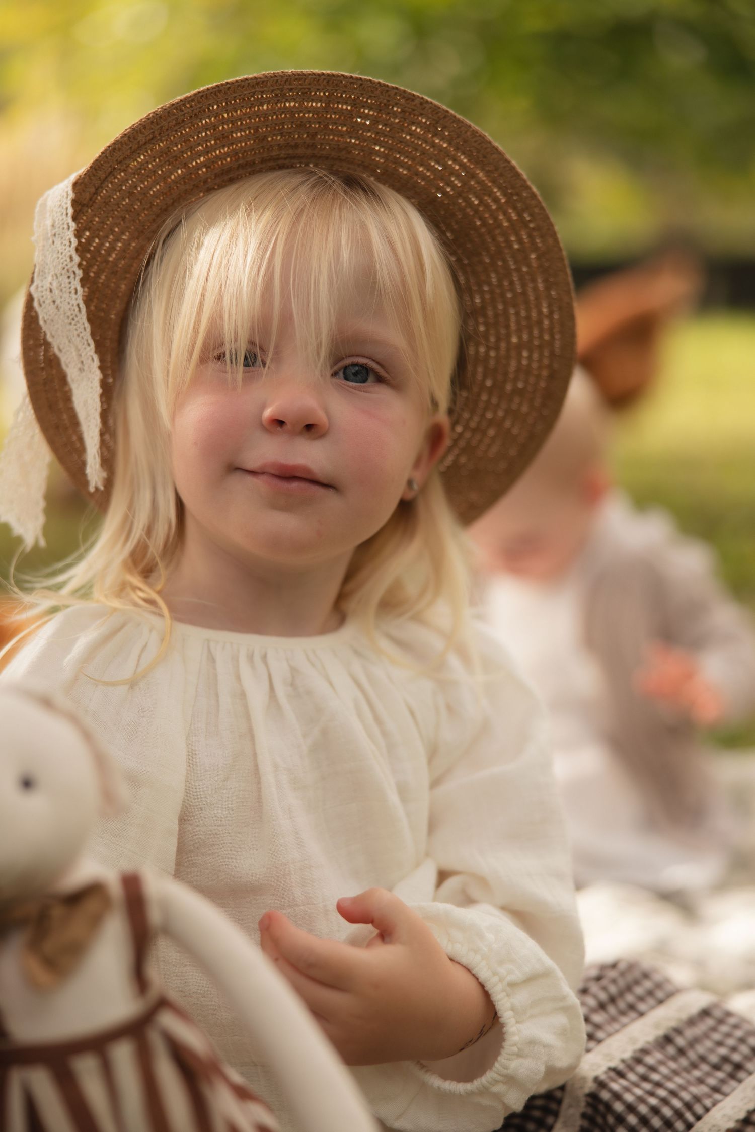 A child wearing a straw hat with lace and white peasant dress strikes a sweet pose outdoors.