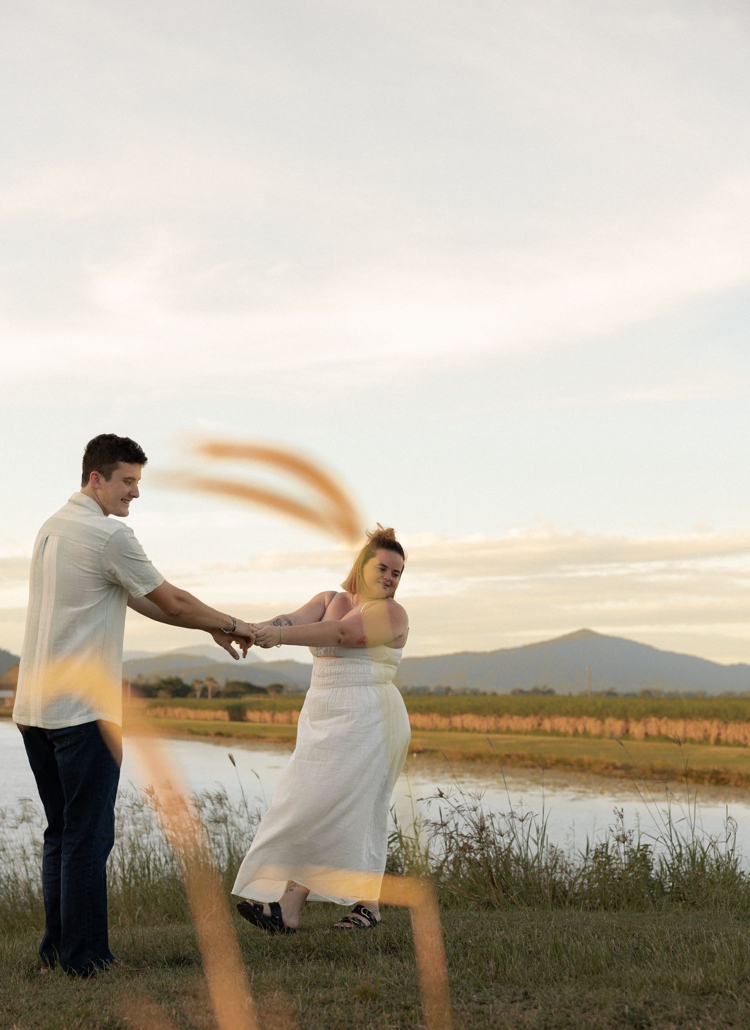 A couple dances together in a white dress and casual attire against a sunset landscape with mountains and fields.