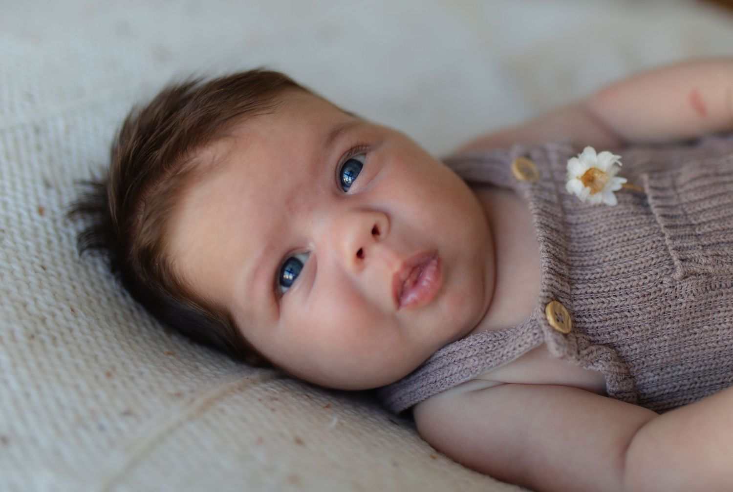 A baby wearing a grey knit outfit with a daisy detail lies on white bedding looking up with bright eyes.