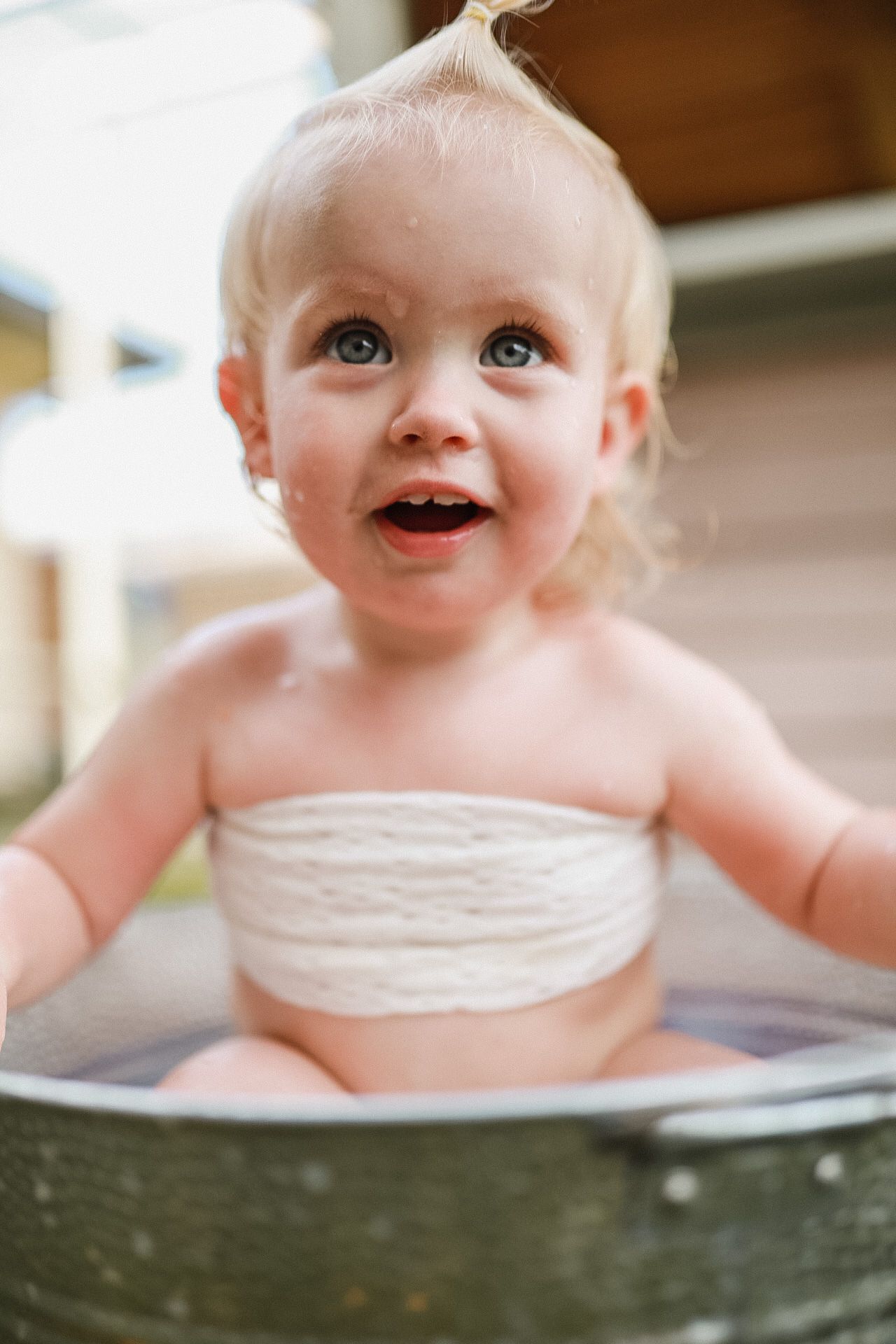 Happy toddler sitting in metal washtub wearing white bandeau top during outdoor playtime.