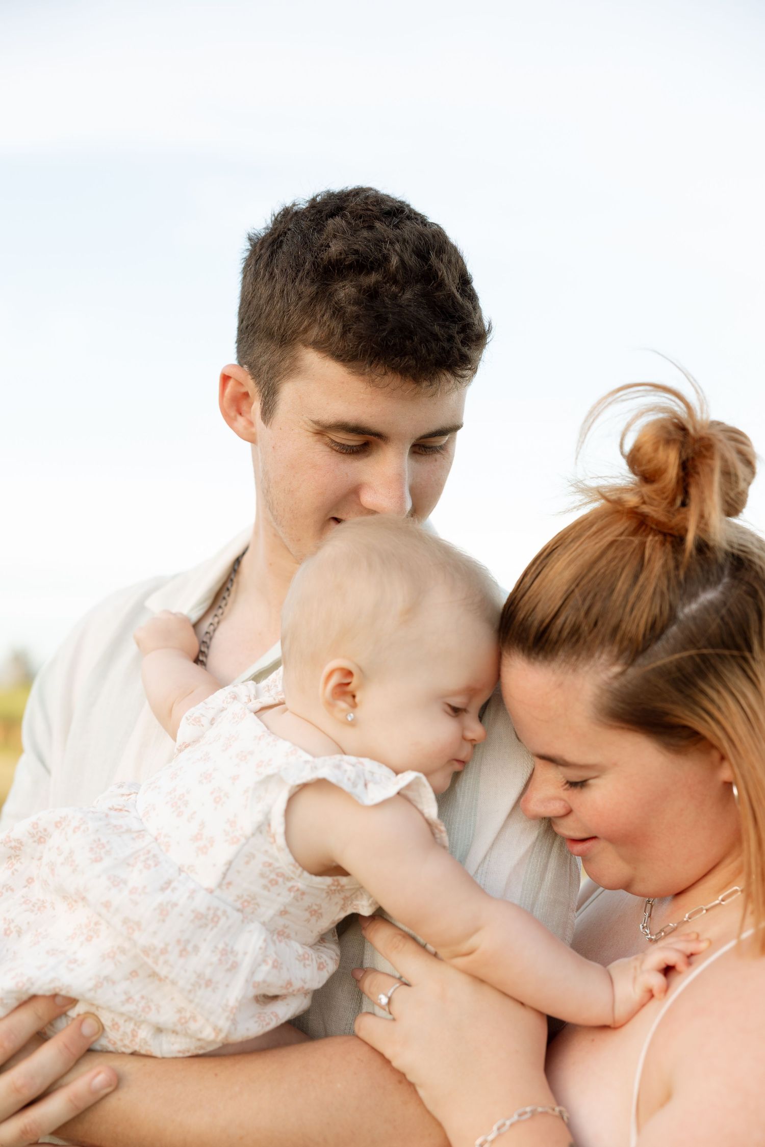 A loving family moment shows tender interactions with their baby wearing a white lace dress in warm sunlight.