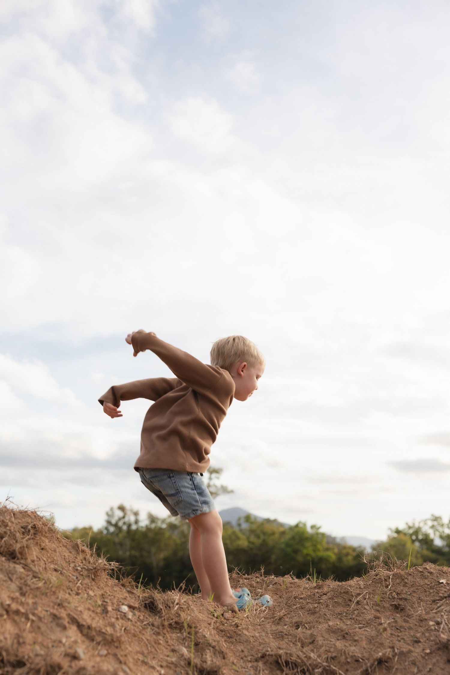A child in brown sweater and shorts balances on dirt mound against cloudy sky.