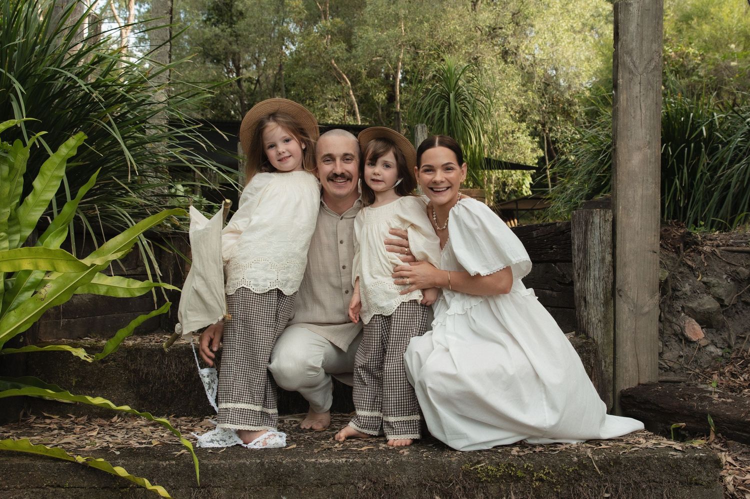Family group in coordinated white and beige outfits sitting together on outdoor stone steps in garden setting.