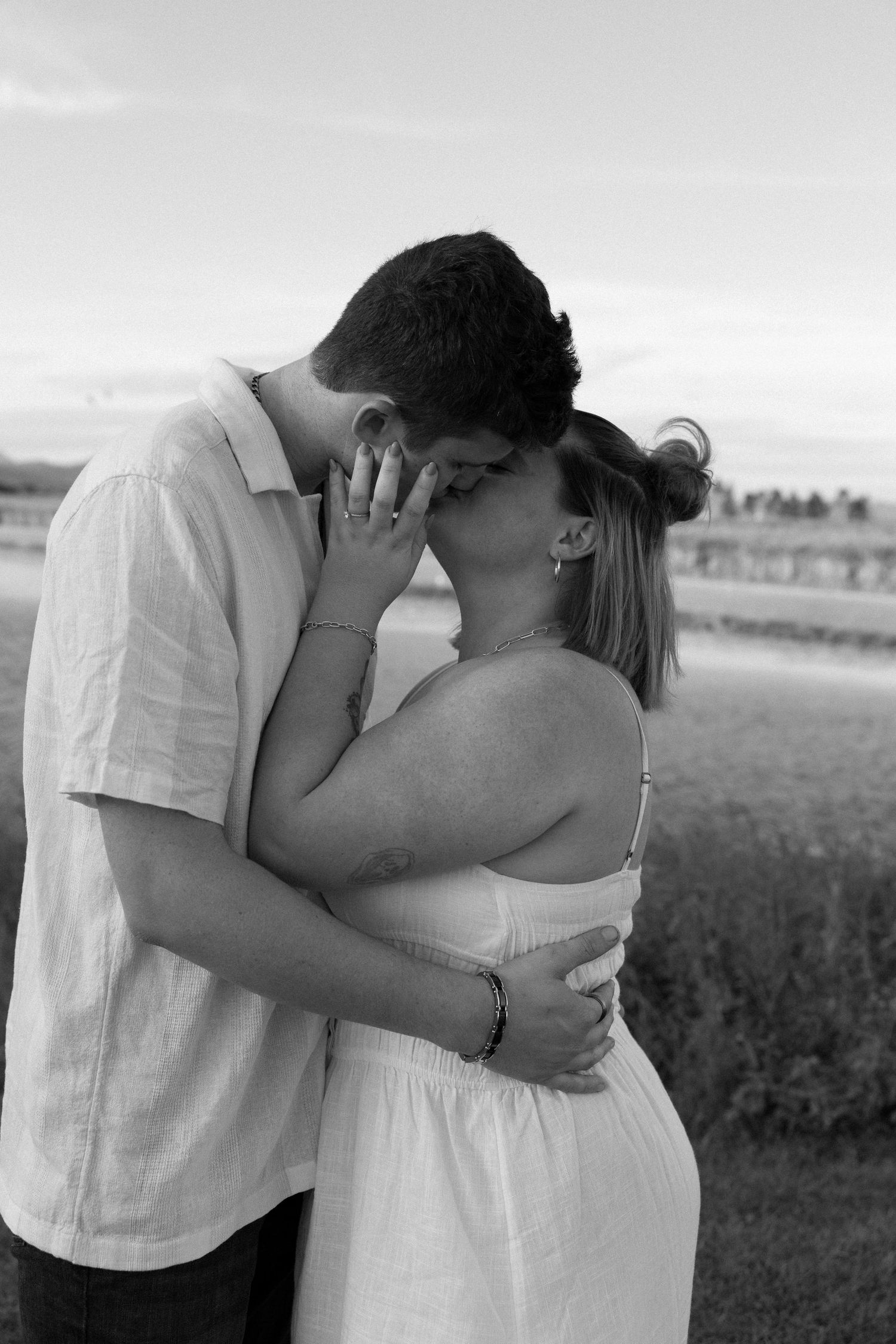 A romantic black and white outdoor photograph of a couple embracing and kissing in a field.