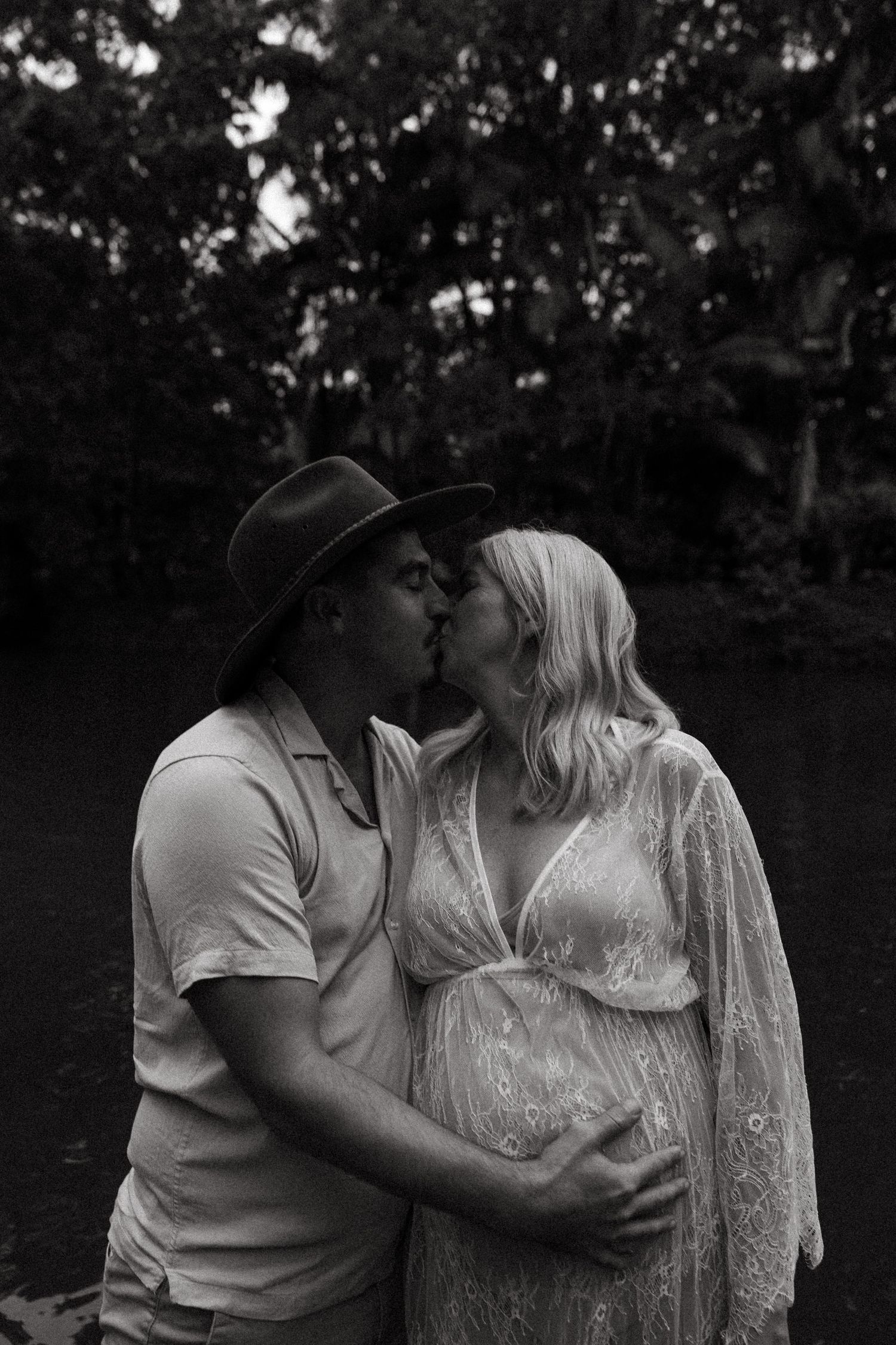 Black and white portrait of a couple embracing outdoors, with a pregnant figure wearing a flowing lace dress.