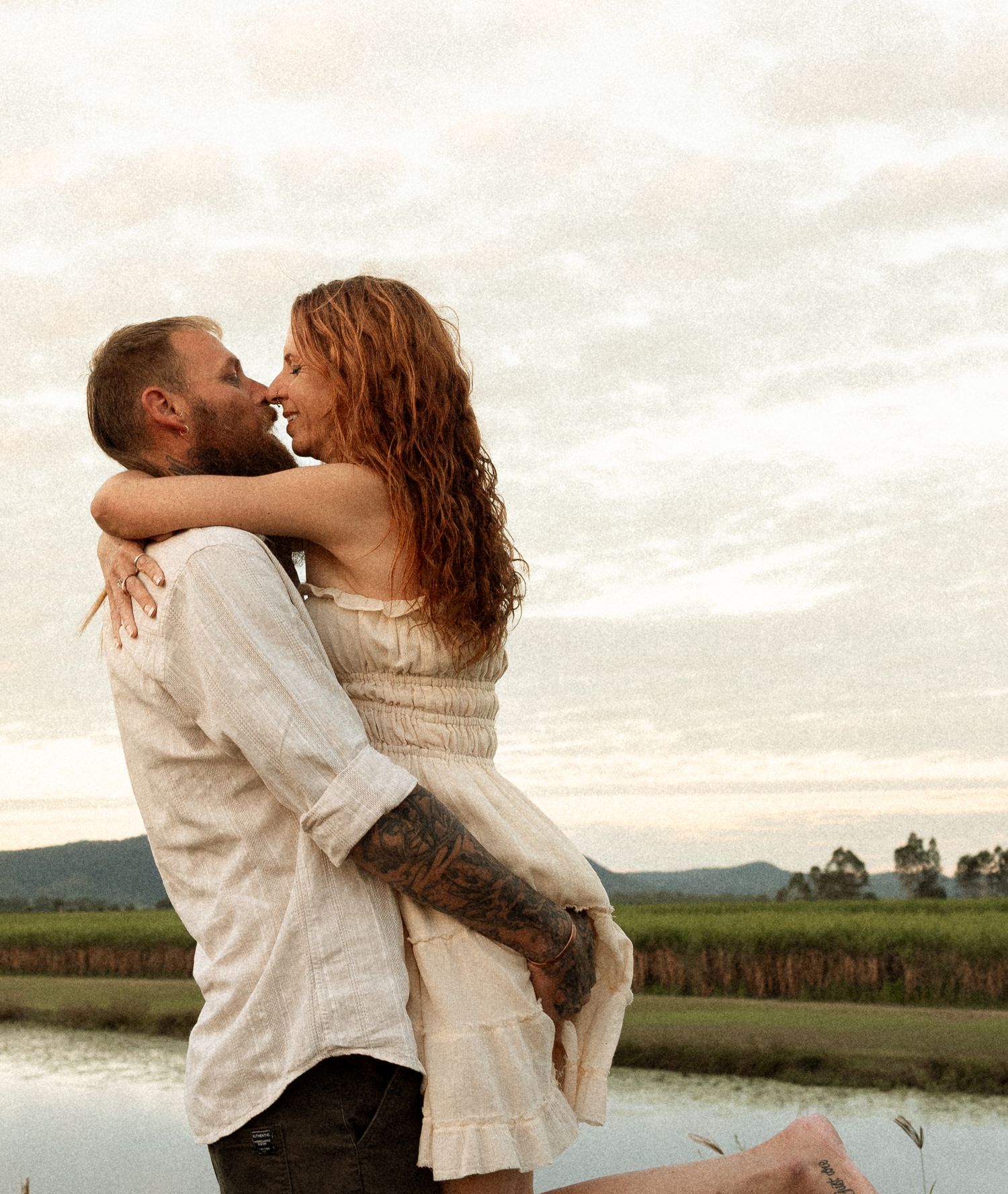 A romantic couple embraces by a serene waterfront with mountains and fields in the background at sunset.