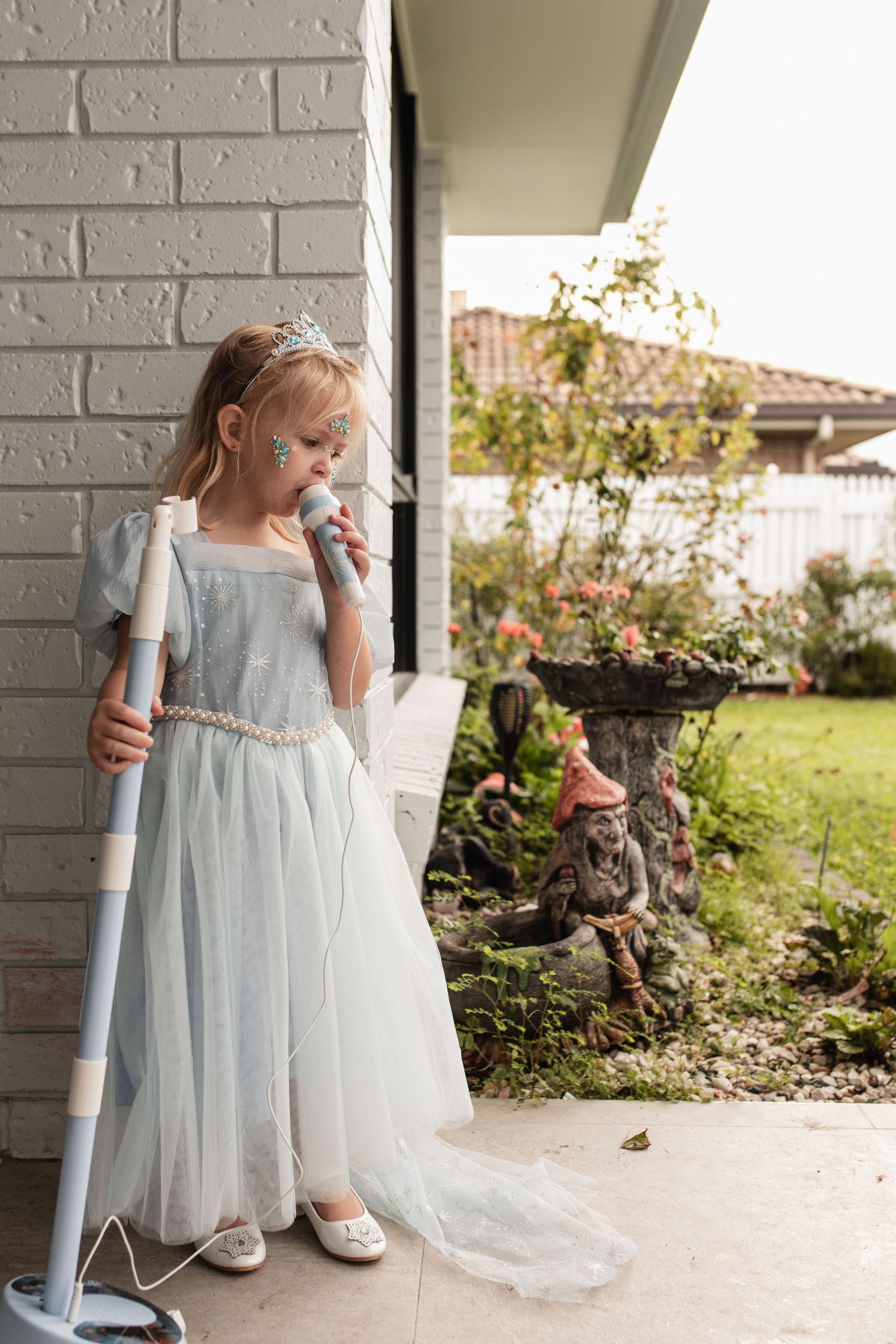 A dreamy scene of a fairy-tale dress and crown against a white brick wall with garden decorations.