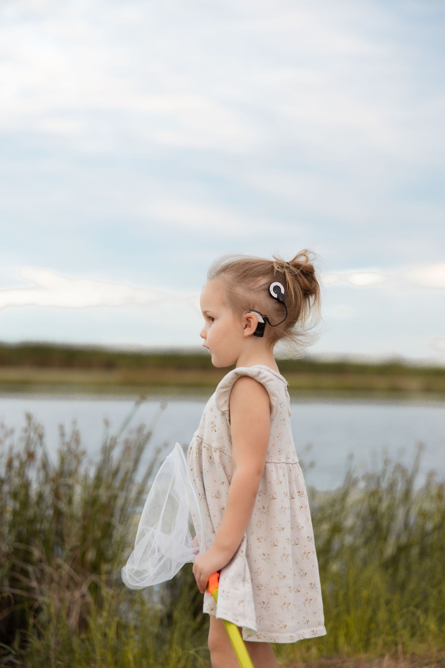 Young child in a light dress holding a butterfly net stands by the waterside at dusk.