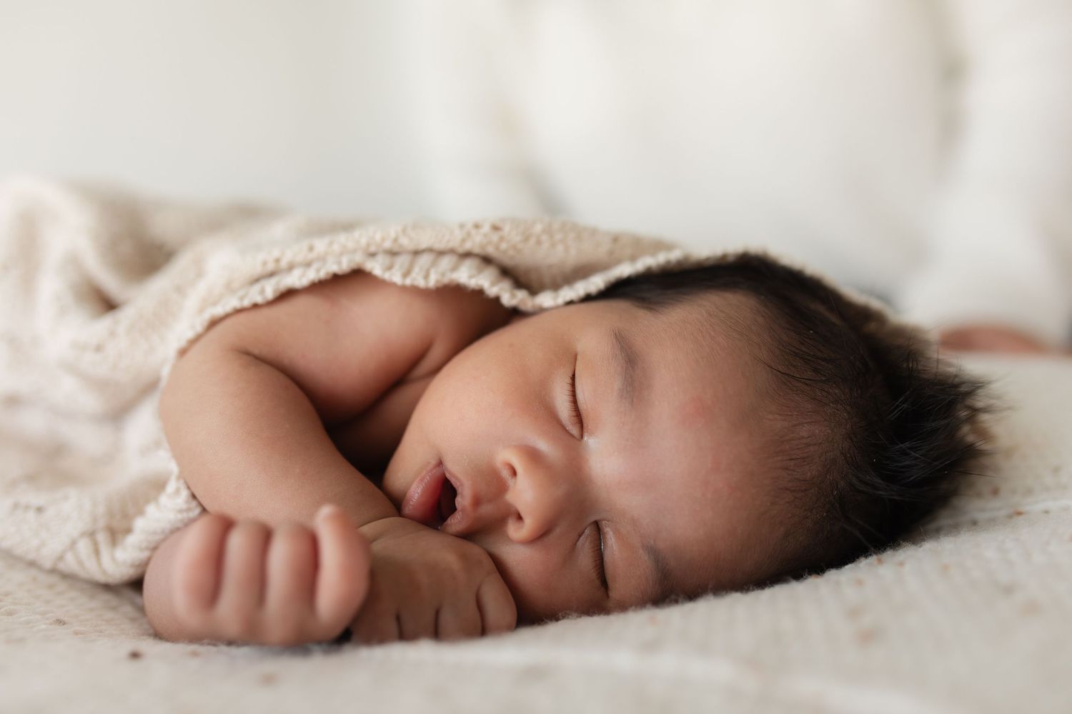 A sleeping newborn baby wrapped in a beige knit blanket rests peacefully on white bedding.