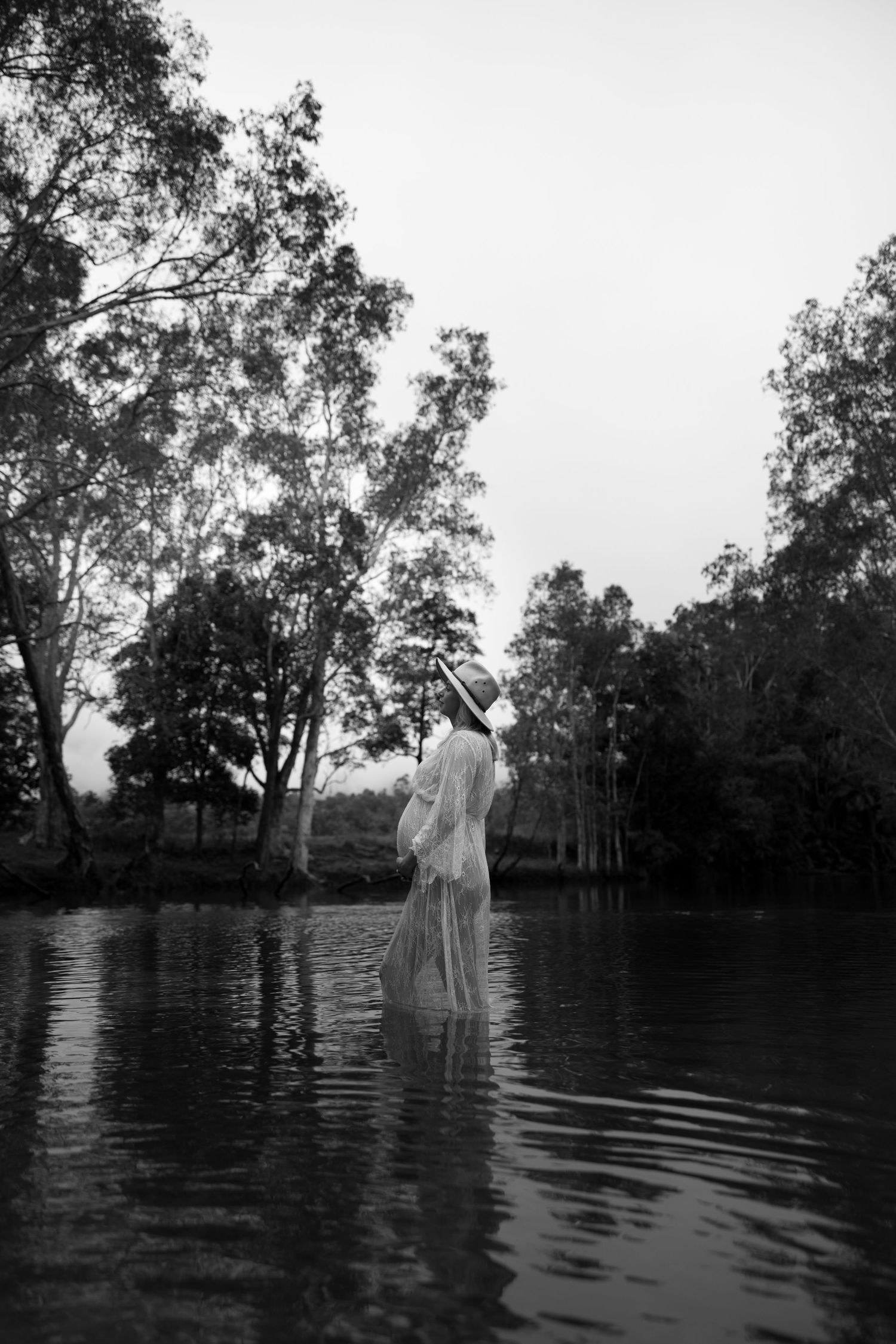 Ethereal figure in white dress standing in still water with tall trees in black and white composition.