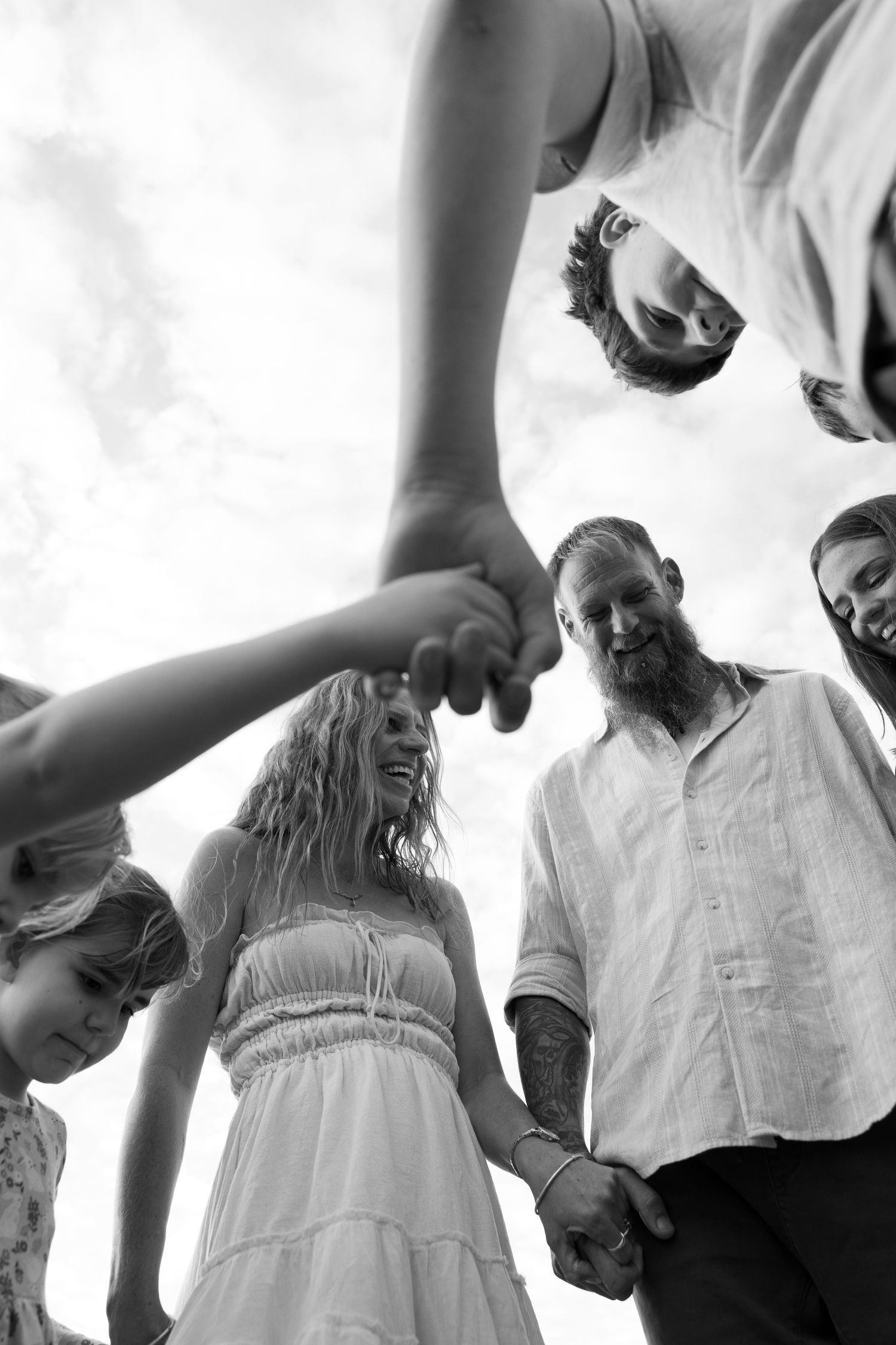 A black and white low-angle view of a group holding hands in a circle formation against a bright sky.