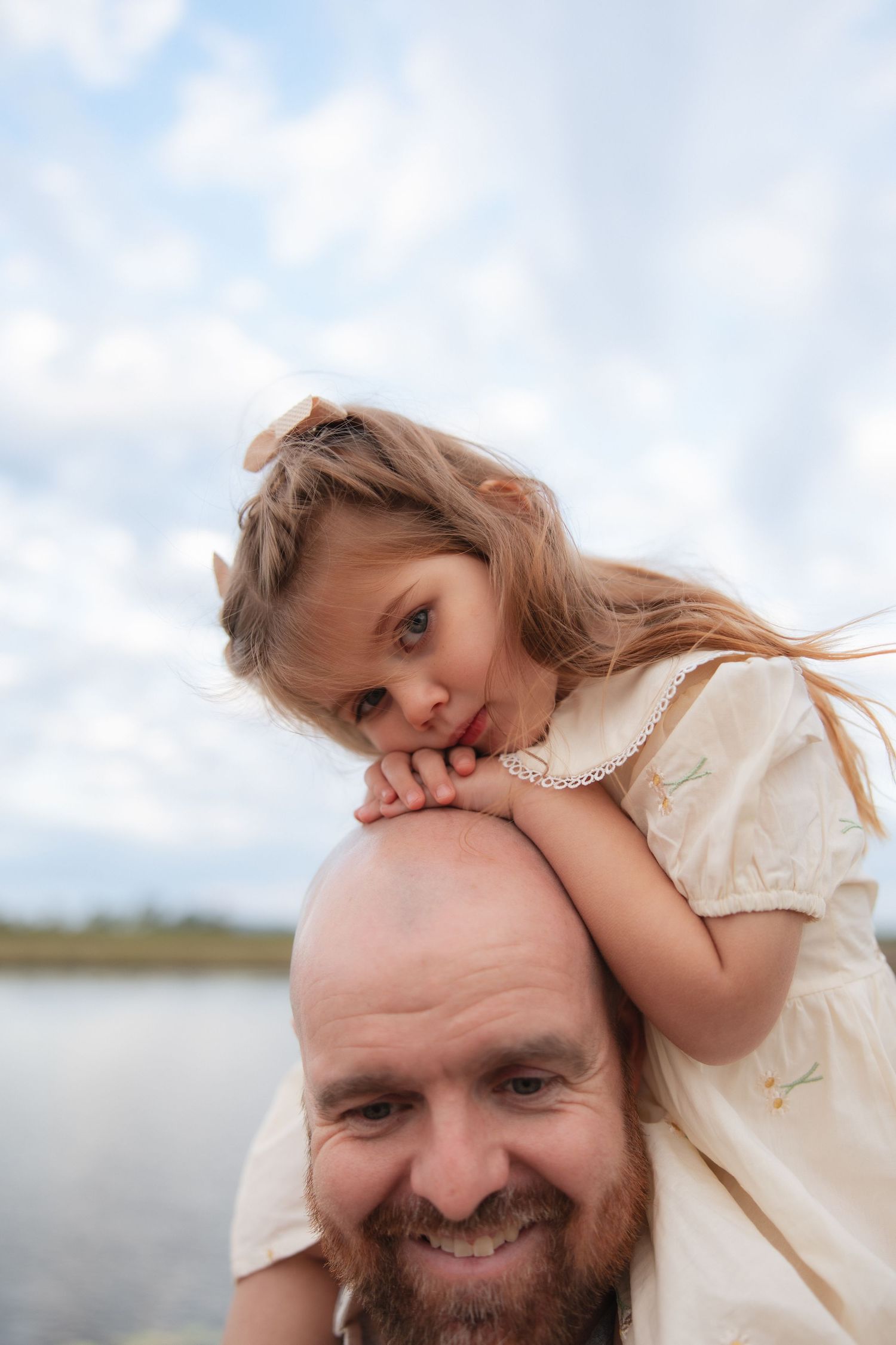 A father and daughter in white clothing share a joyful moment together near a waterfront on a cloudy day.