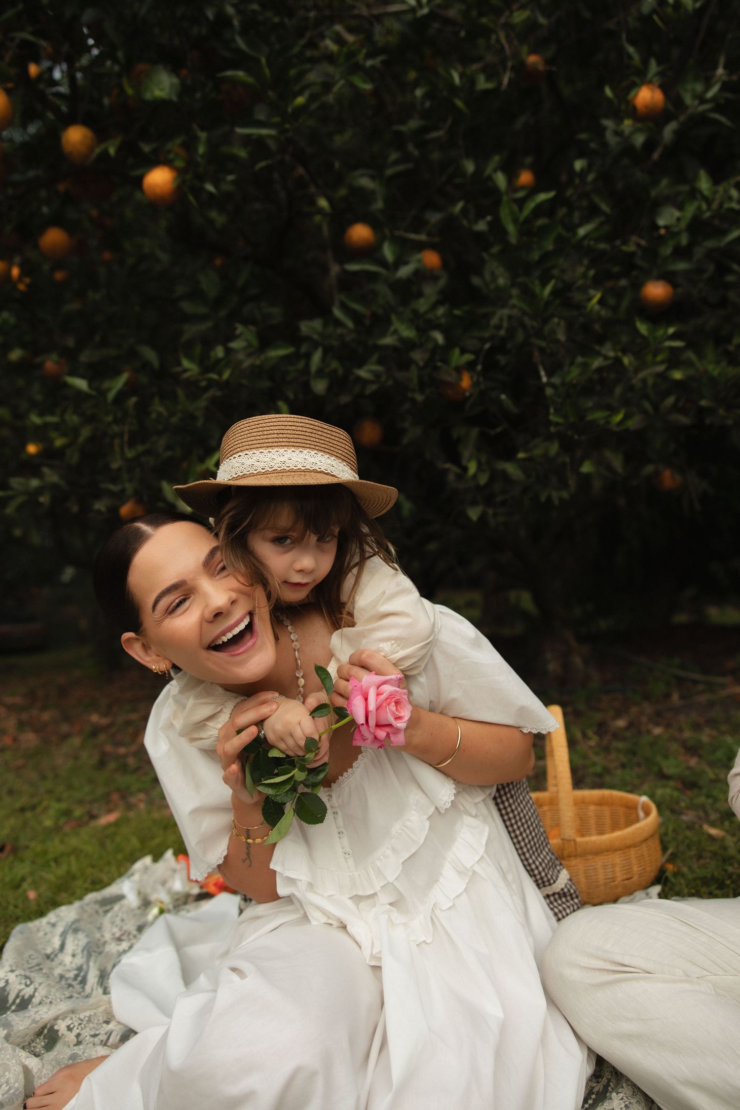 Two friends in white vintage dresses laugh together during a picnic in an orange grove on a sunny afternoon.