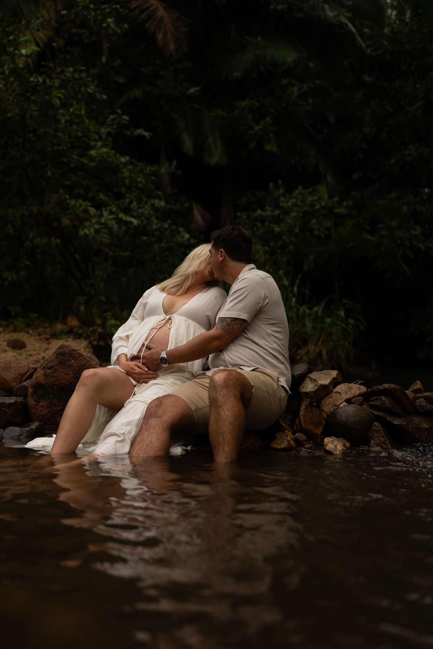 A romantic couple shares an intimate moment sitting in shallow water surrounded by rocks and lush greenery.