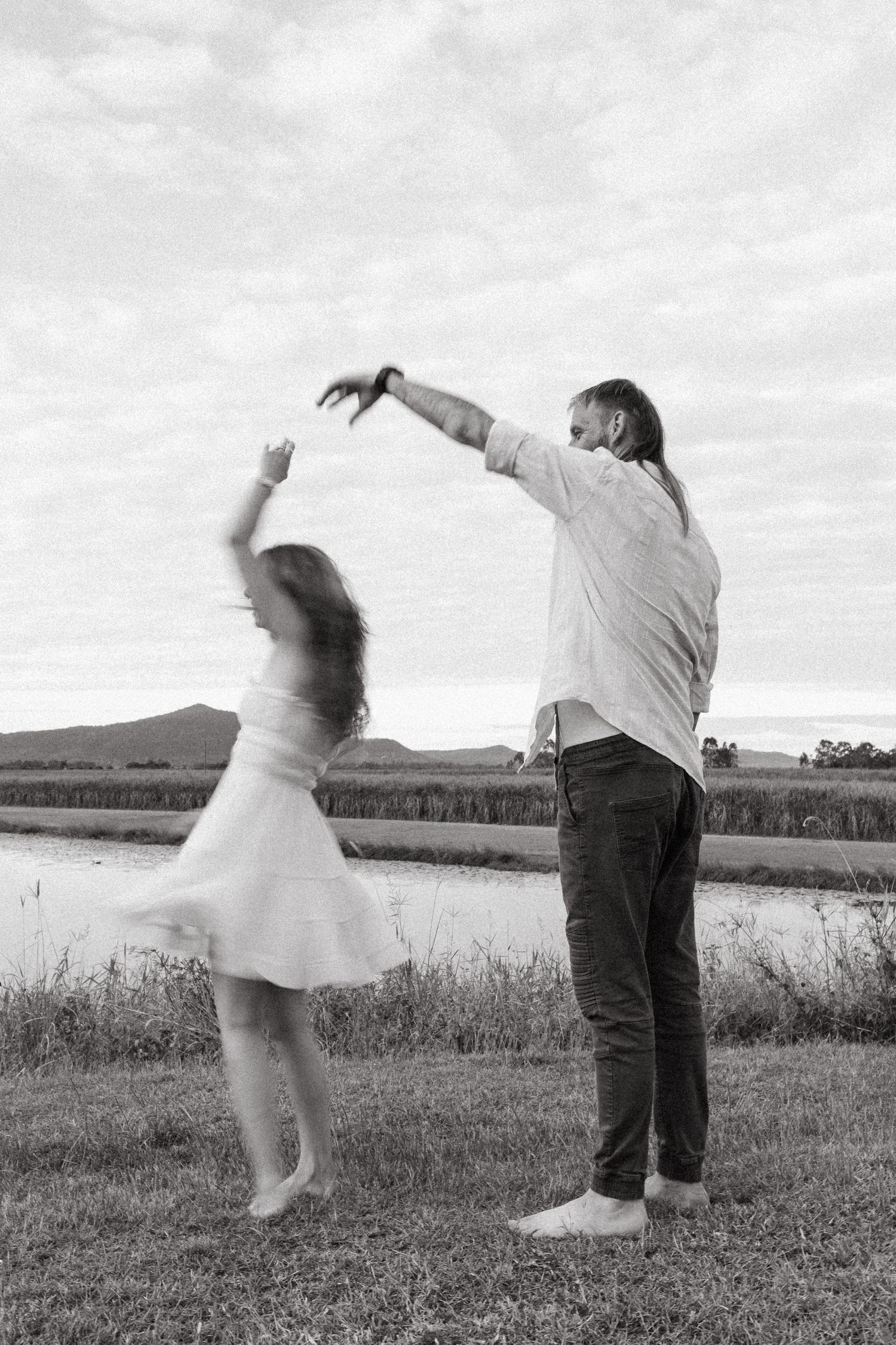 A couple dancing joyfully together in a field against a cloudy sky, captured in black and white.
