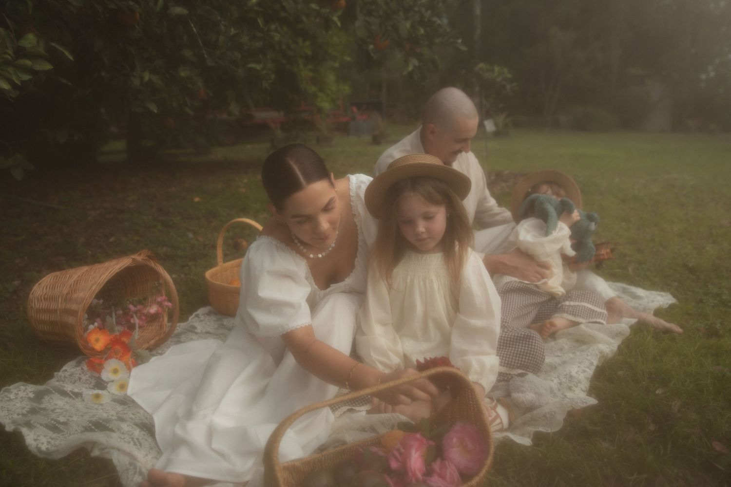 A family shares an intimate outdoor moment on a lace blanket in a garden setting with dappled sunlight.