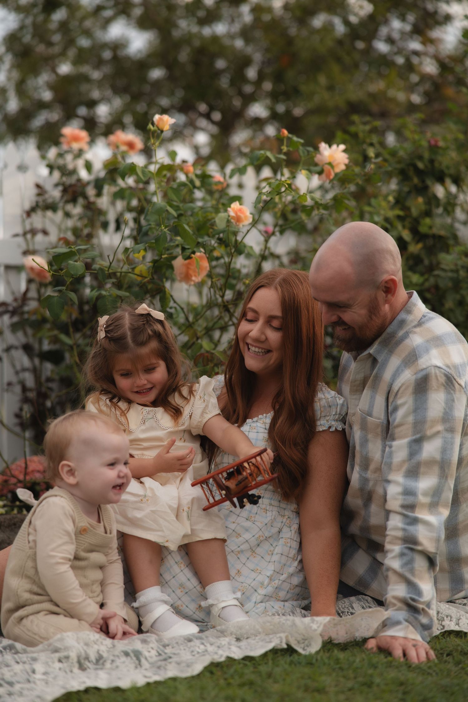 Family sits together on white blanket in garden with blooming peach roses in background during sunset.