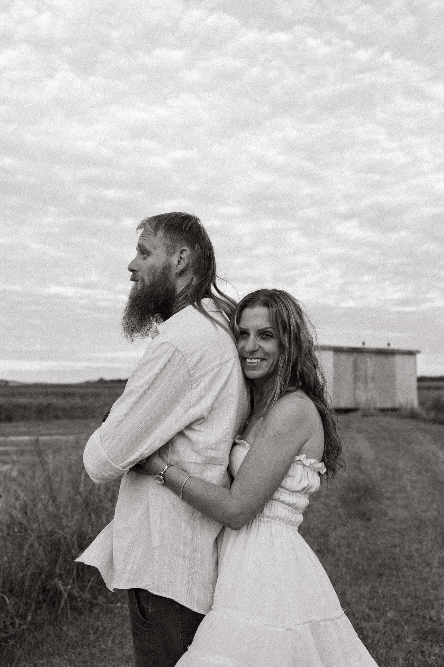 A black and white portrait of a couple in white clothing embracing outdoors with a rustic structure in background.