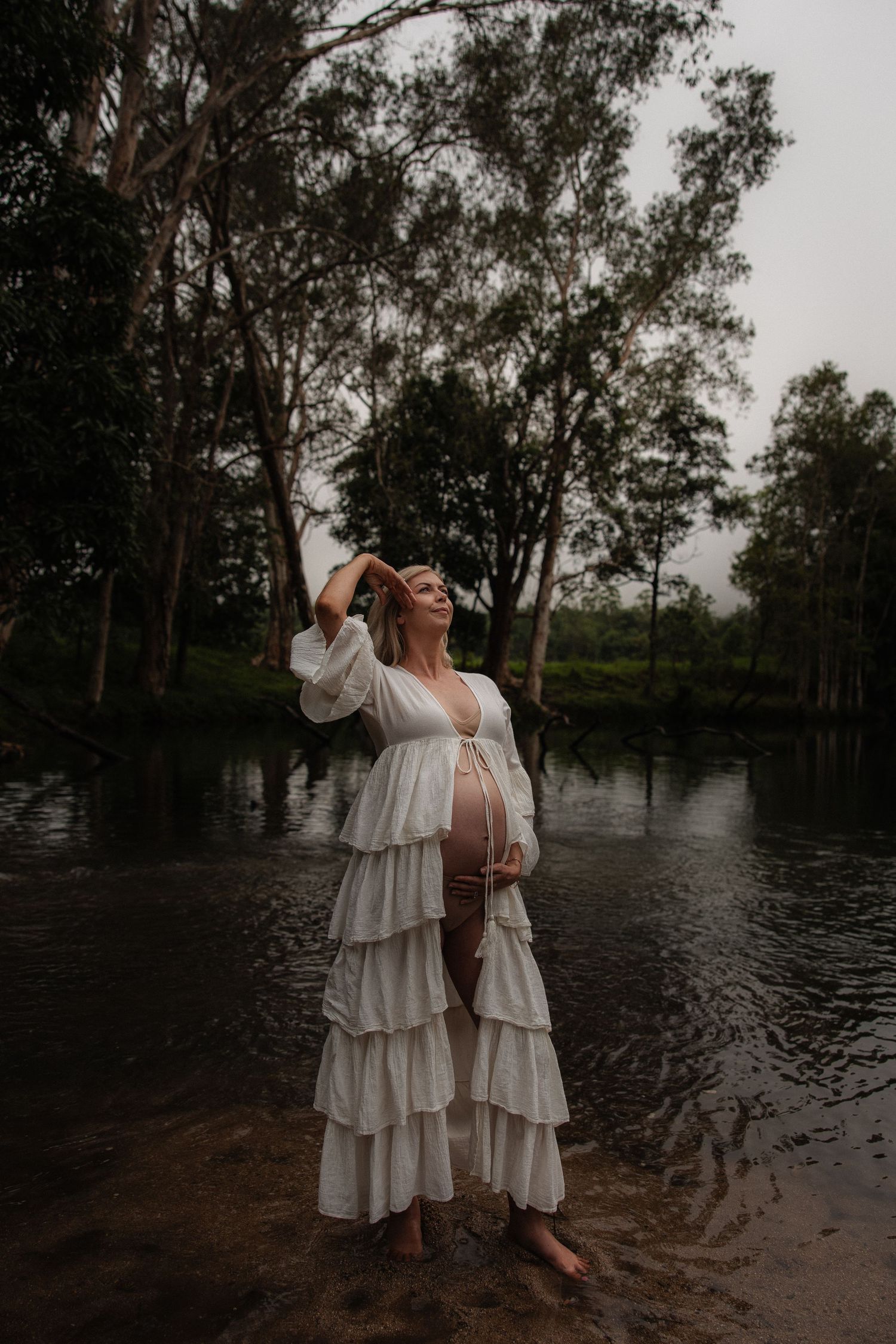 A person in a flowing white tiered dress stands barefoot by a river during a moody maternity photoshoot.
